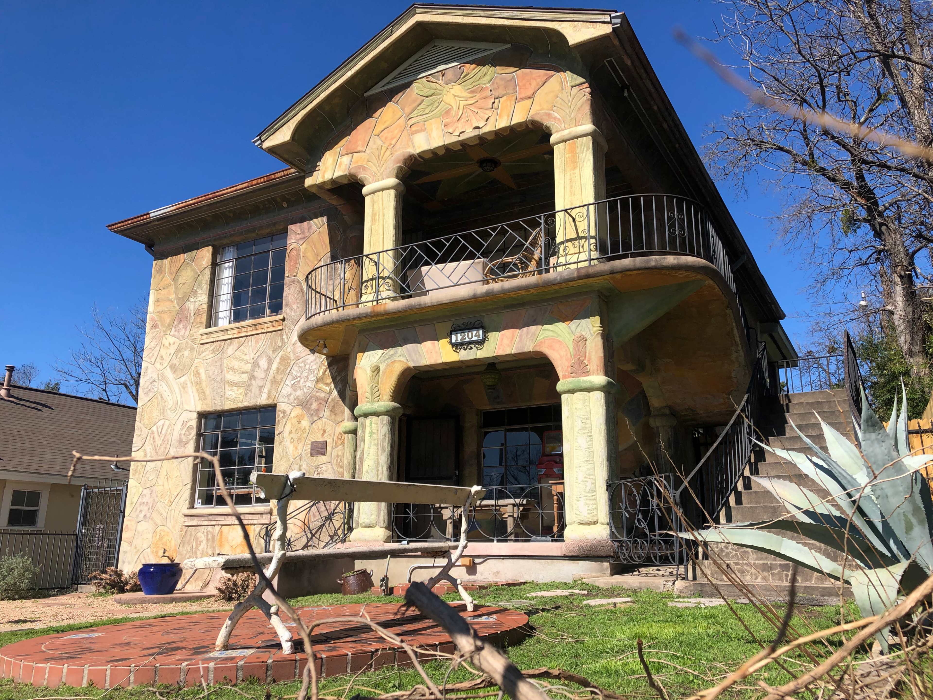 The image shows a two-story house with colorful mural-like decorations on its exterior, featuring a balcony supported by decorative columns and surrounded by a garden.