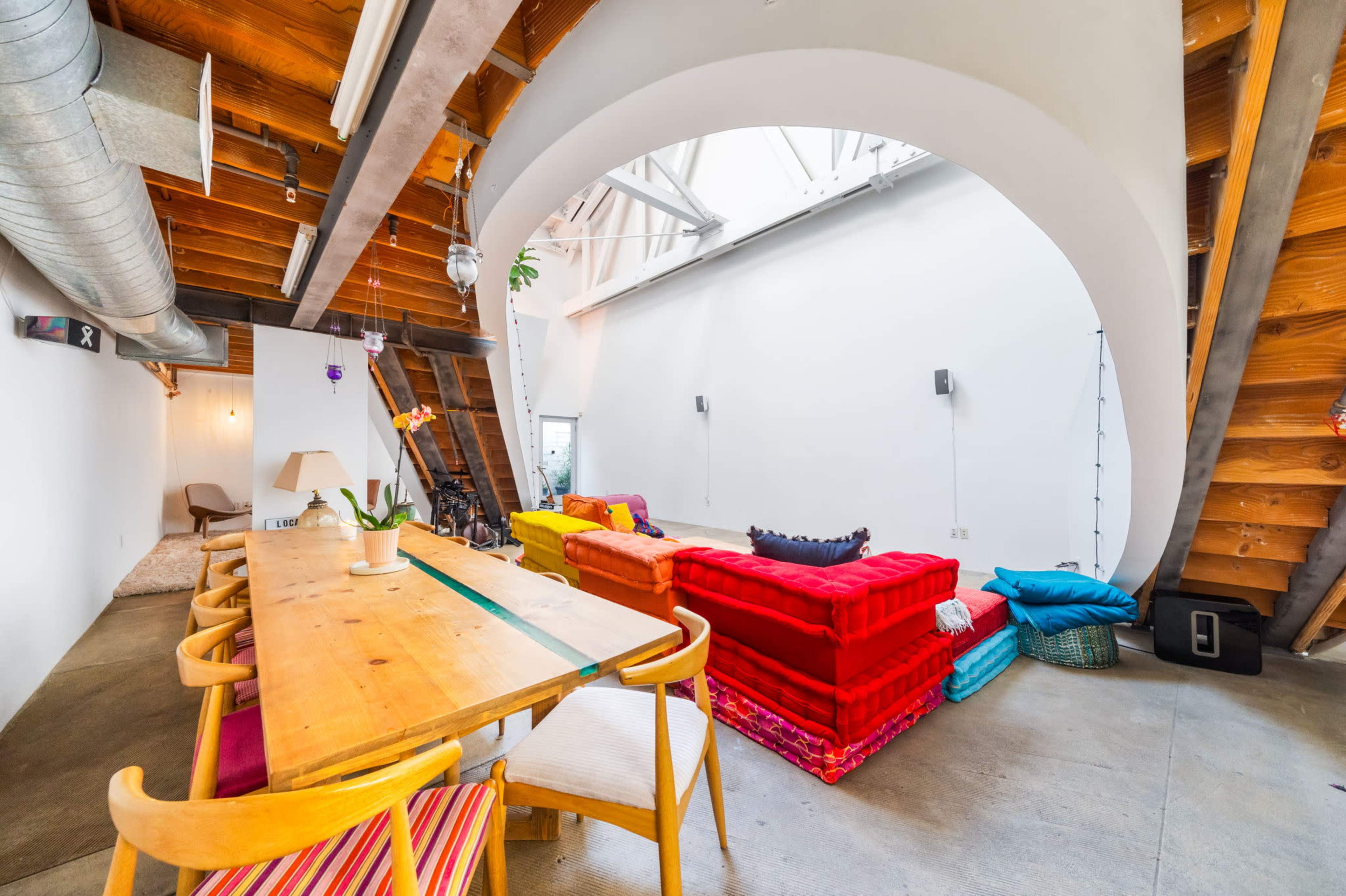 A spacious living area with a wooden dining table, colorful sofas arranged in a semi-enclosed space, and a high ceiling with exposed beams.