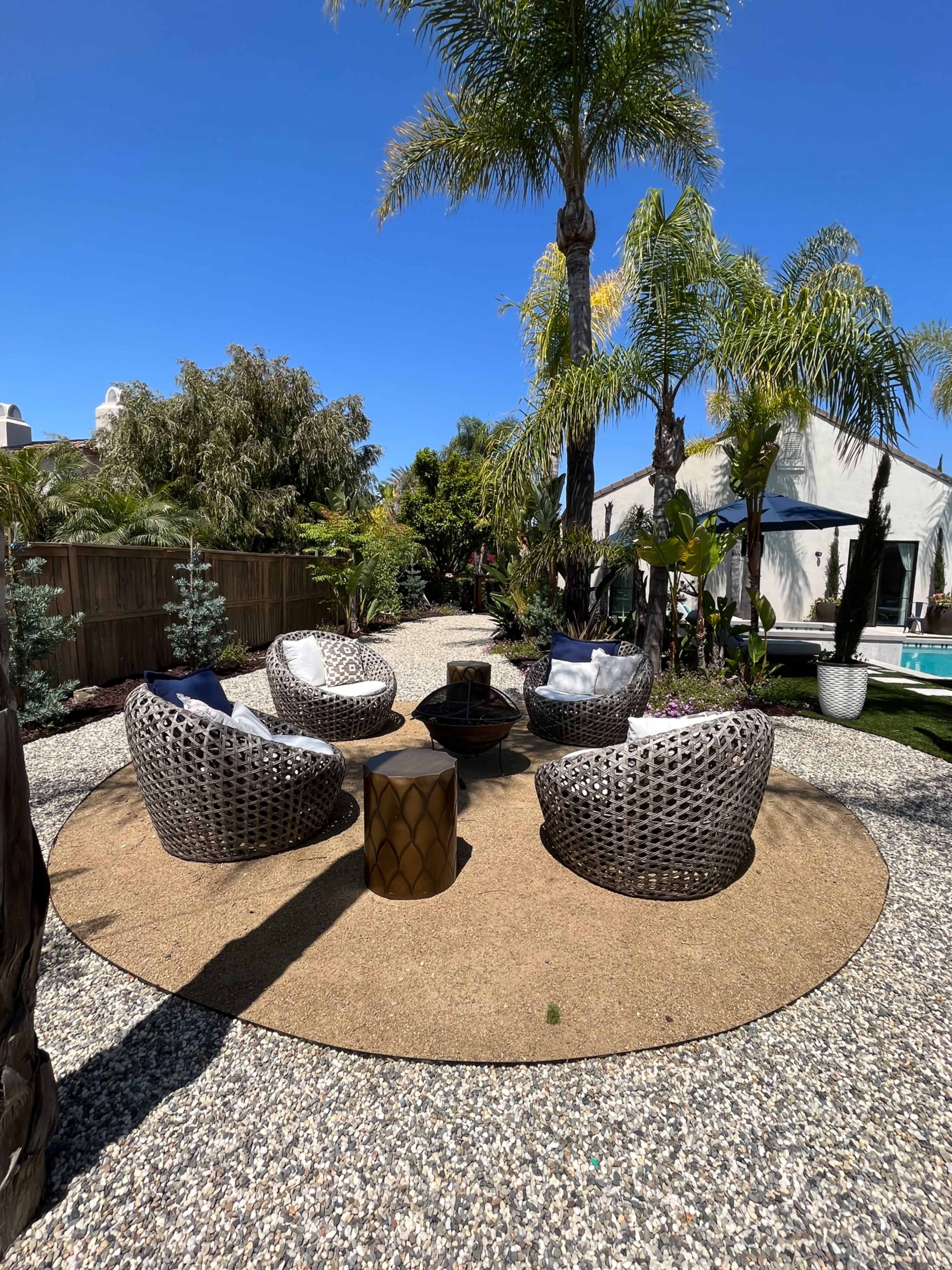 A circular seating area made up of four woven chairs surrounding a central fire pit is set amidst a landscaped garden with palm trees and gravel.
