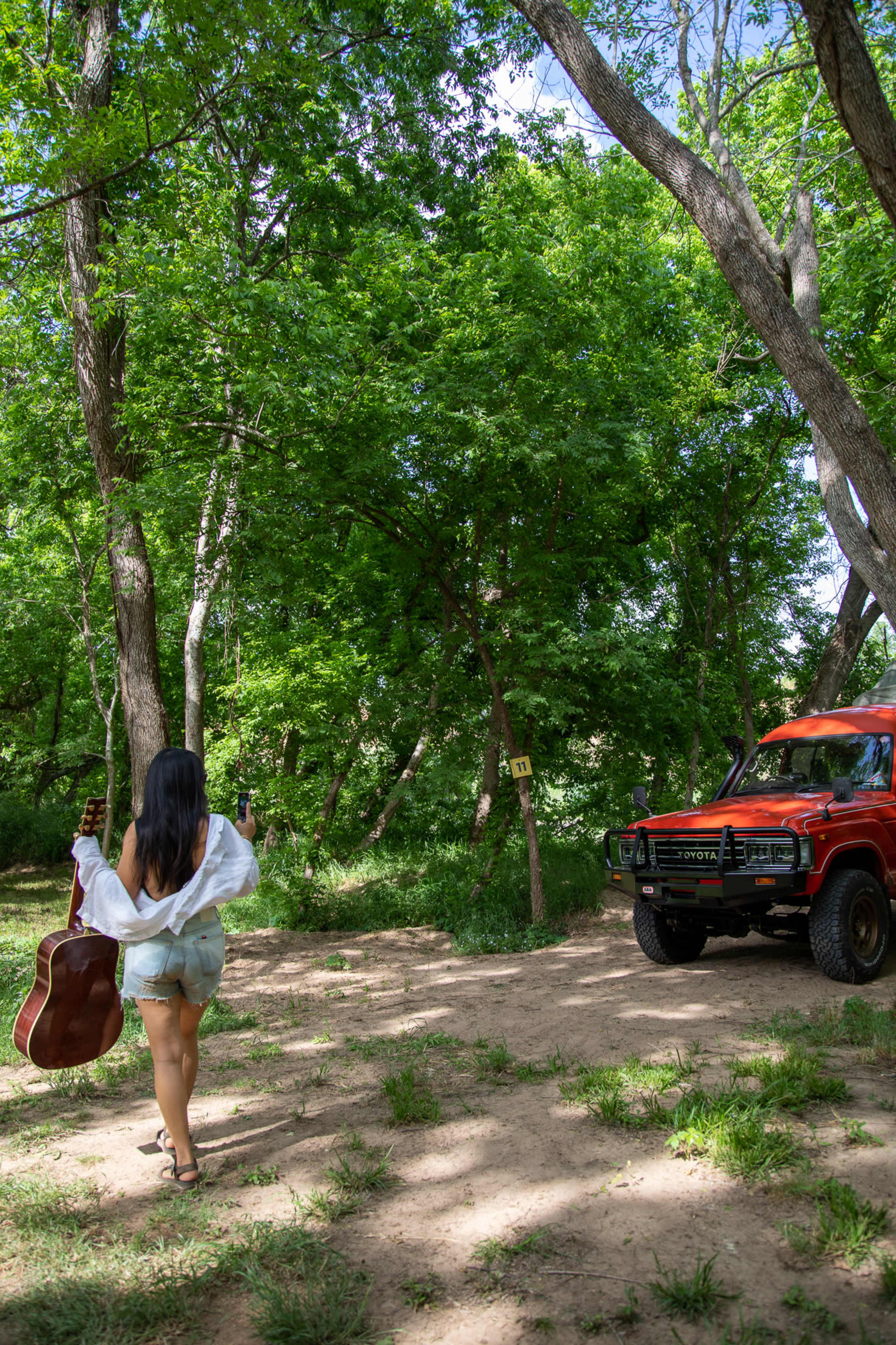 A person walks towards a red truck while carrying a guitar in a wooded area.