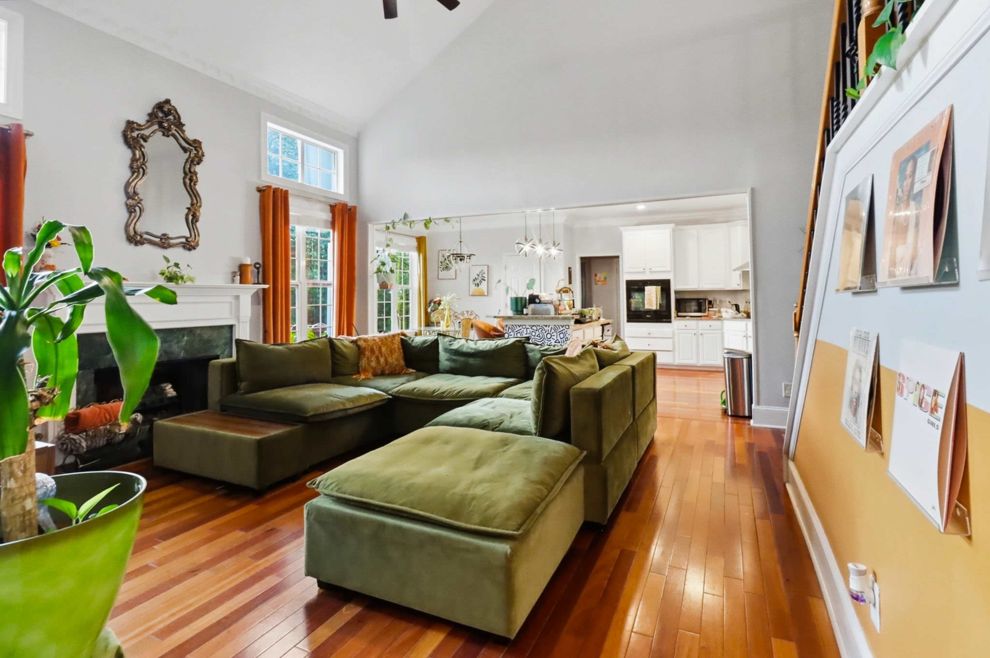 The image depicts a spacious living room with a large green sectional sofa, hardwood flooring, and a view of an adjoining kitchen through an open layout.