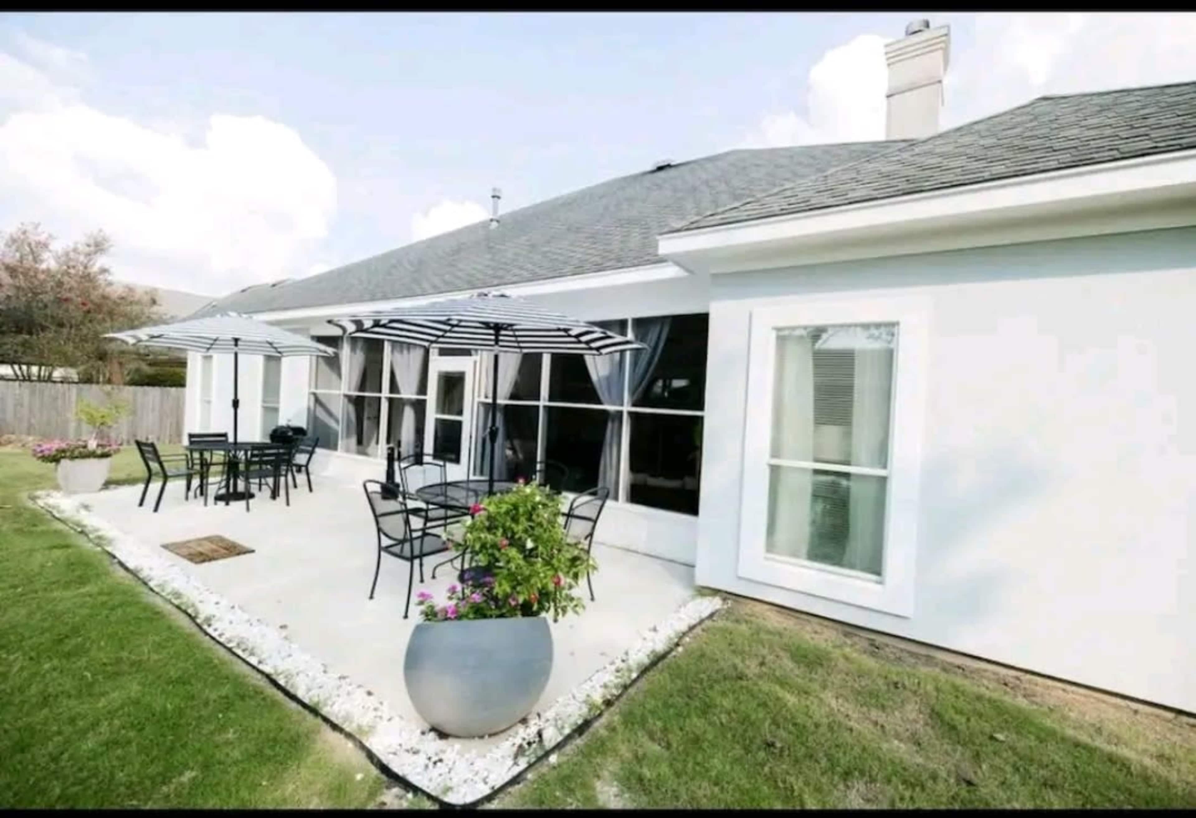 A modern patio area with metal furniture, large umbrellas, and flowerpots, adjacent to a house with large windows.