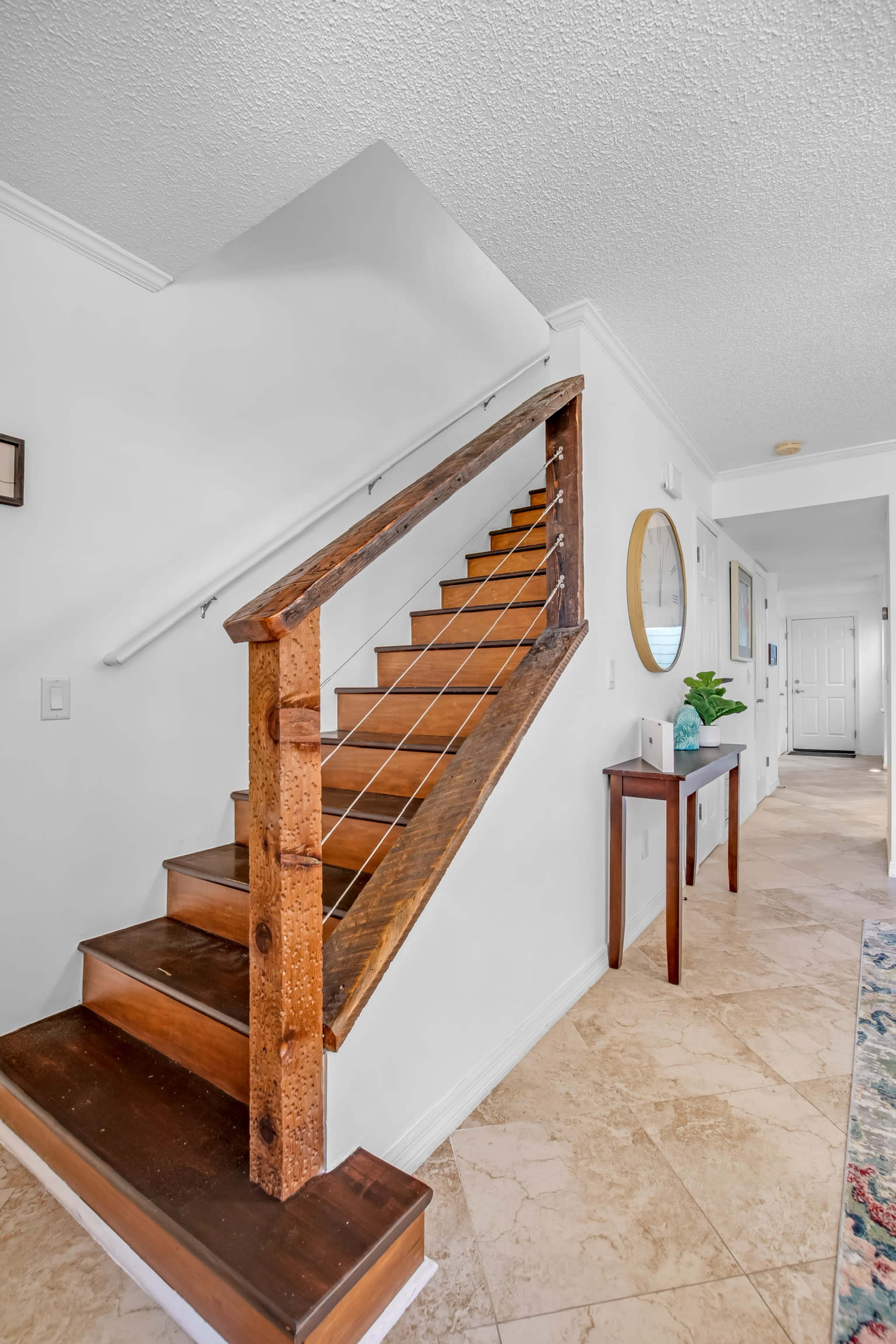 A well-lit hallway featuring a wooden staircase with a modern railing, leading to an upper level, and a console table beside a round mirror.