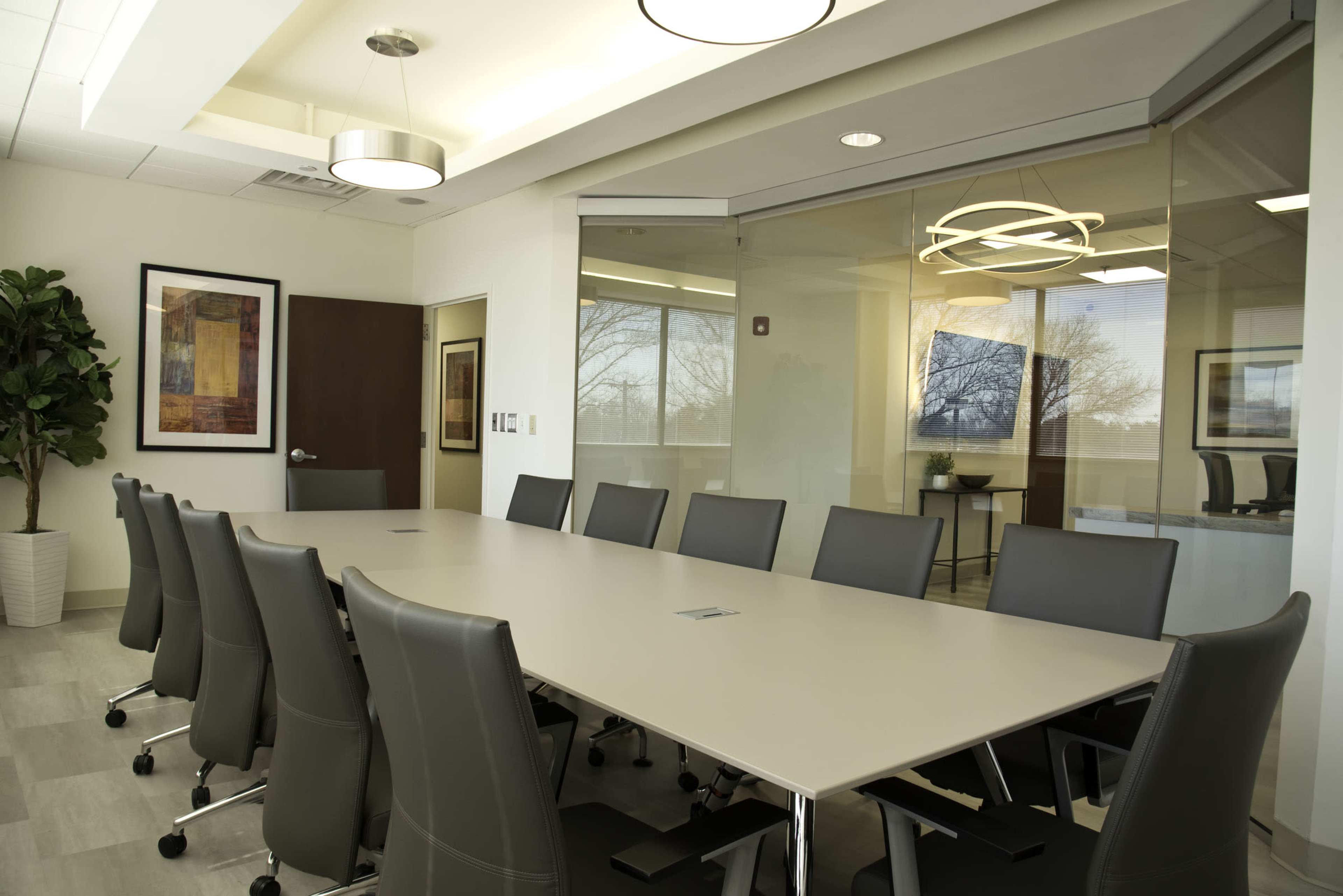 A modern conference room features a large rectangular table surrounded by ergonomic chairs and a glass wall offering a view of an outside landscape.