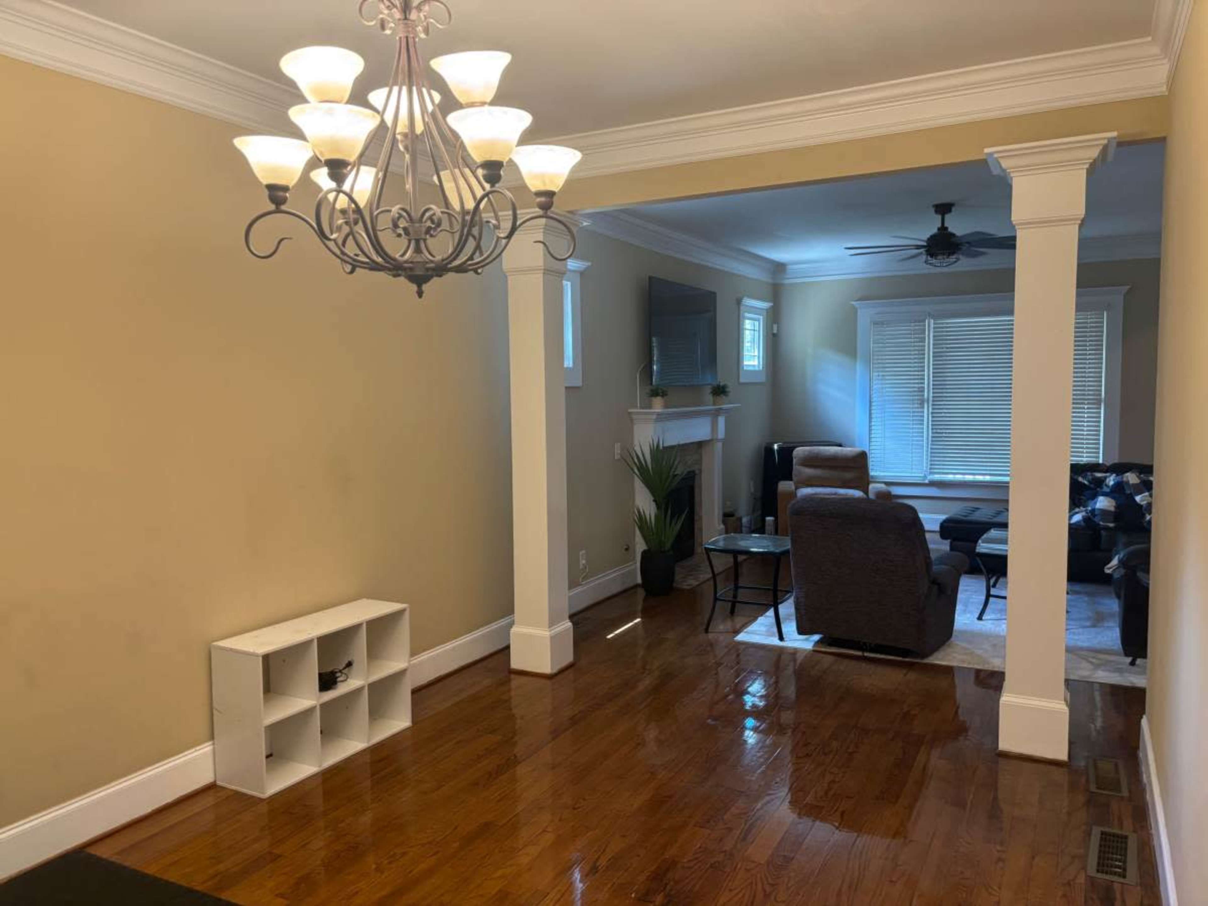 The image shows a living room with wooden flooring, a chandelier, a small shelving unit, and furniture visible in a connected space.