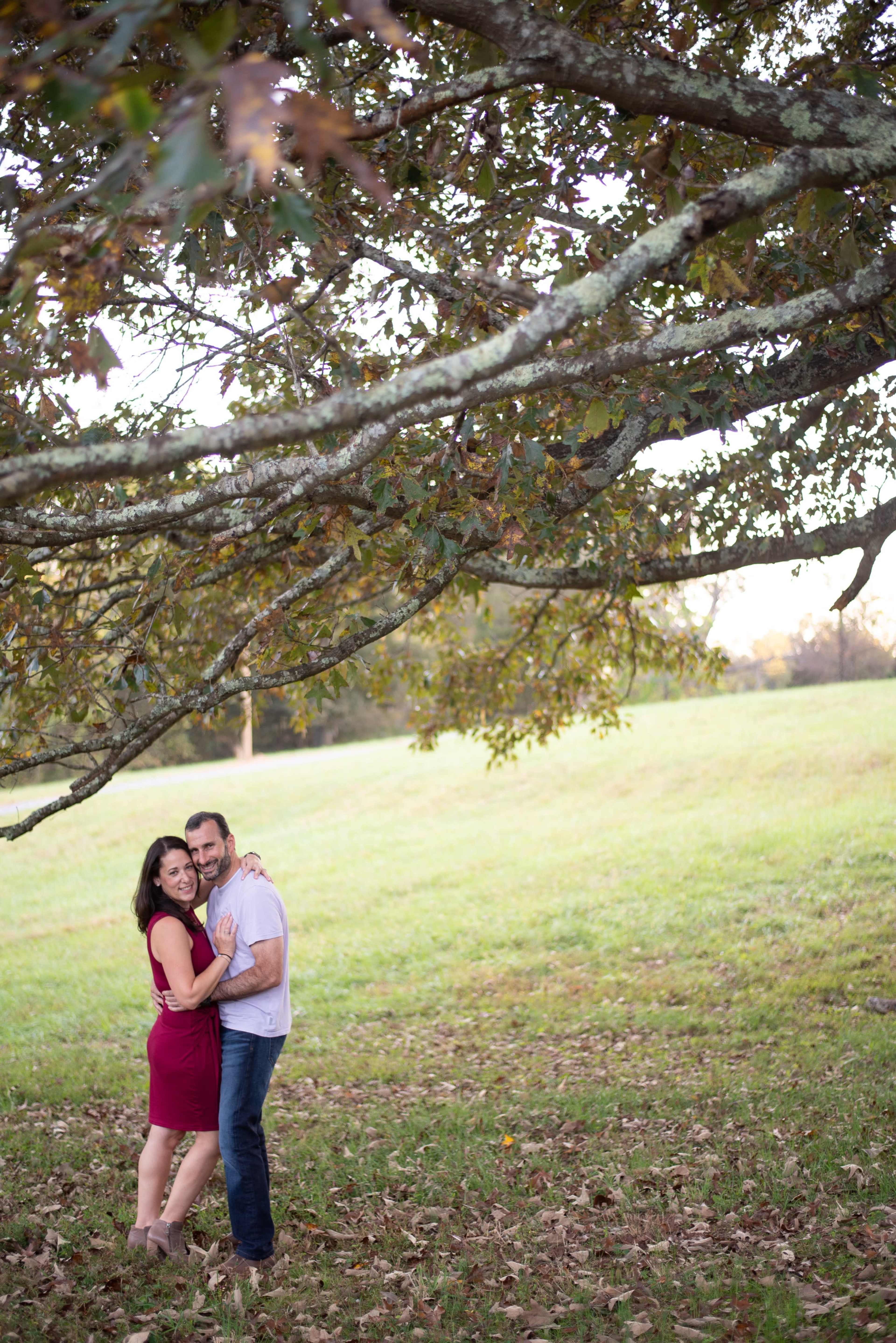 A couple stands embraced under a tree with a grassy field in the background.