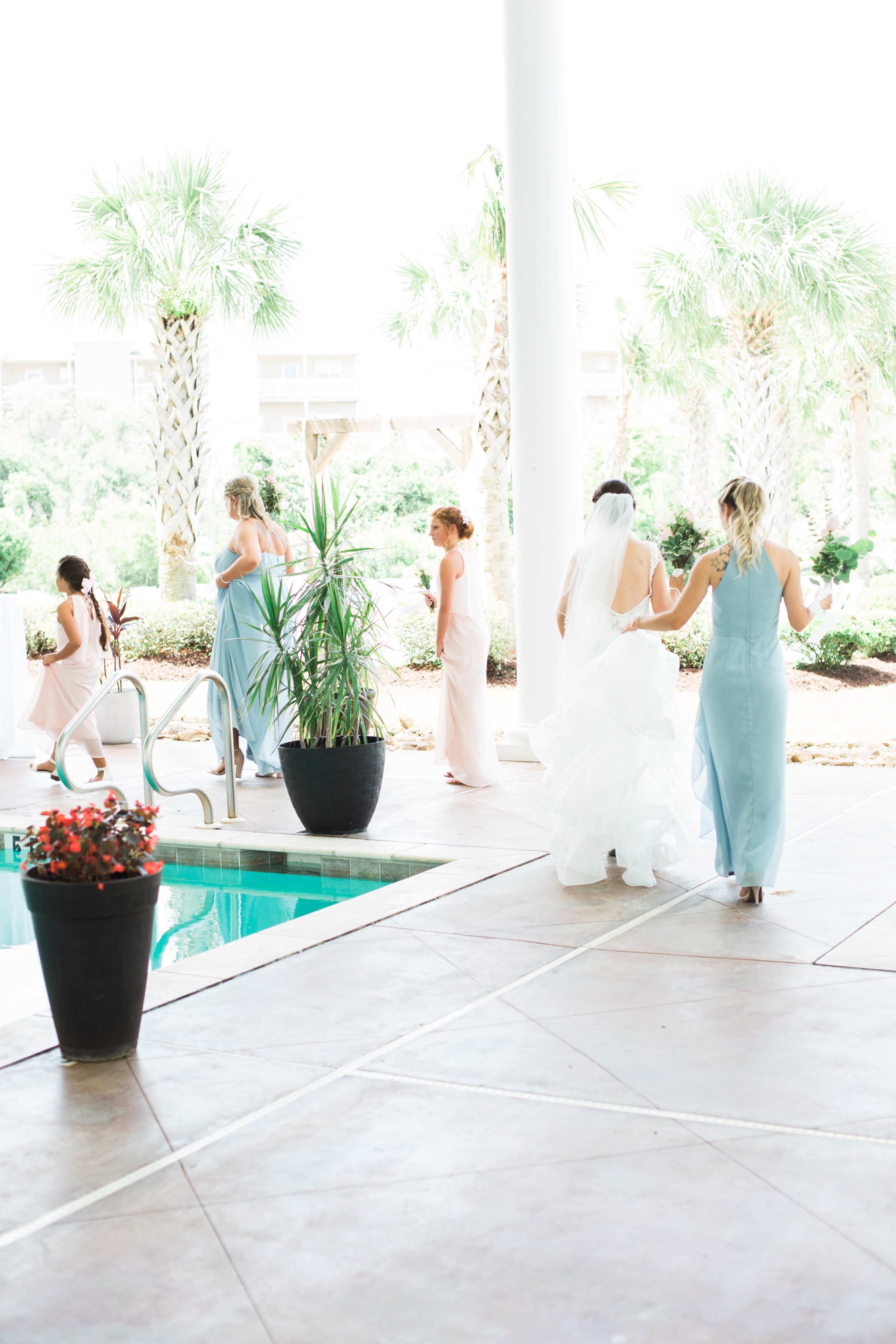 A group of women in formal dresses walks near a pool area surrounded by palm trees.
