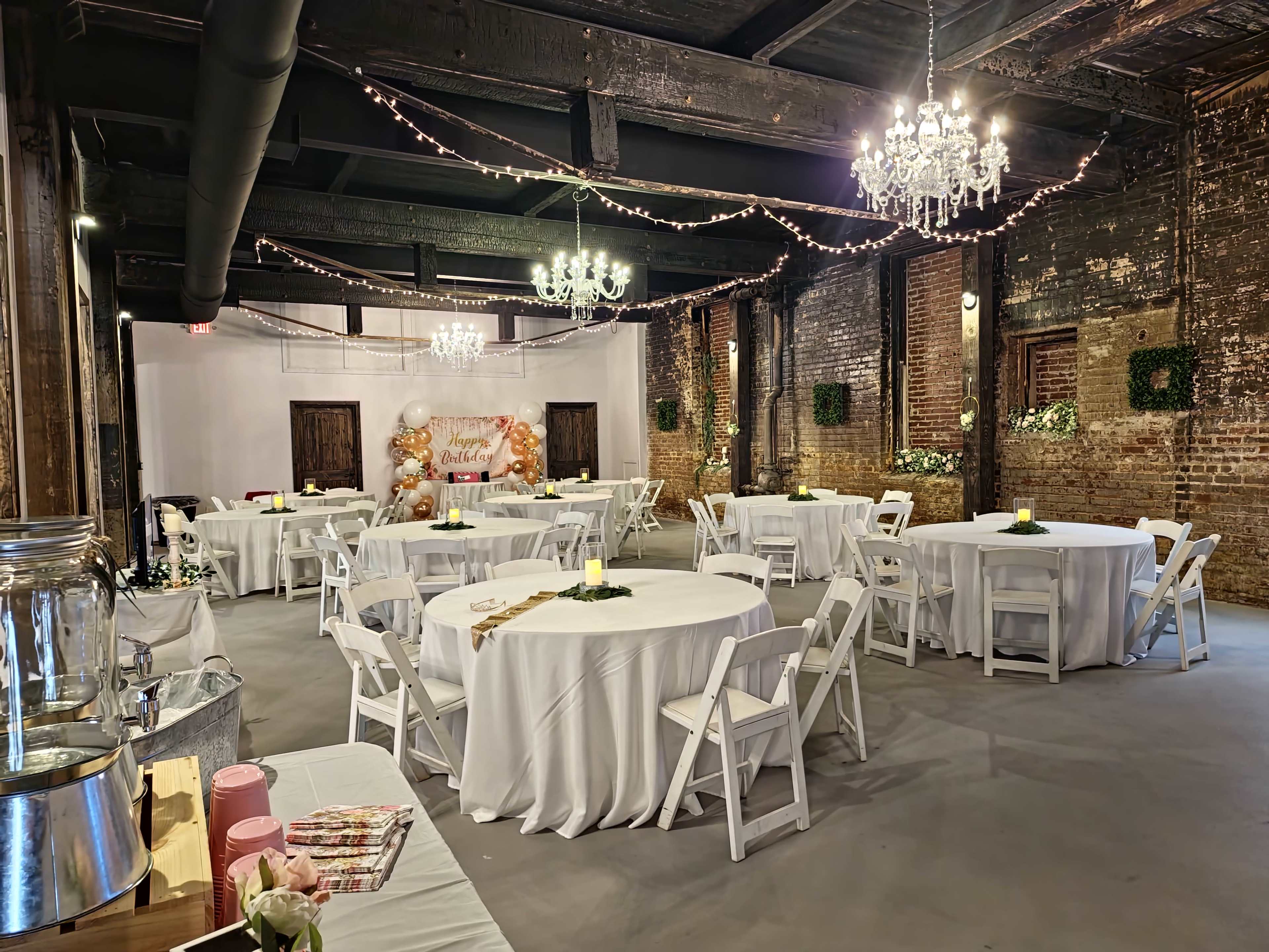 A decorated event space features round tables with white tablecloths, string lights overhead, and chandeliers, set against exposed brick walls.