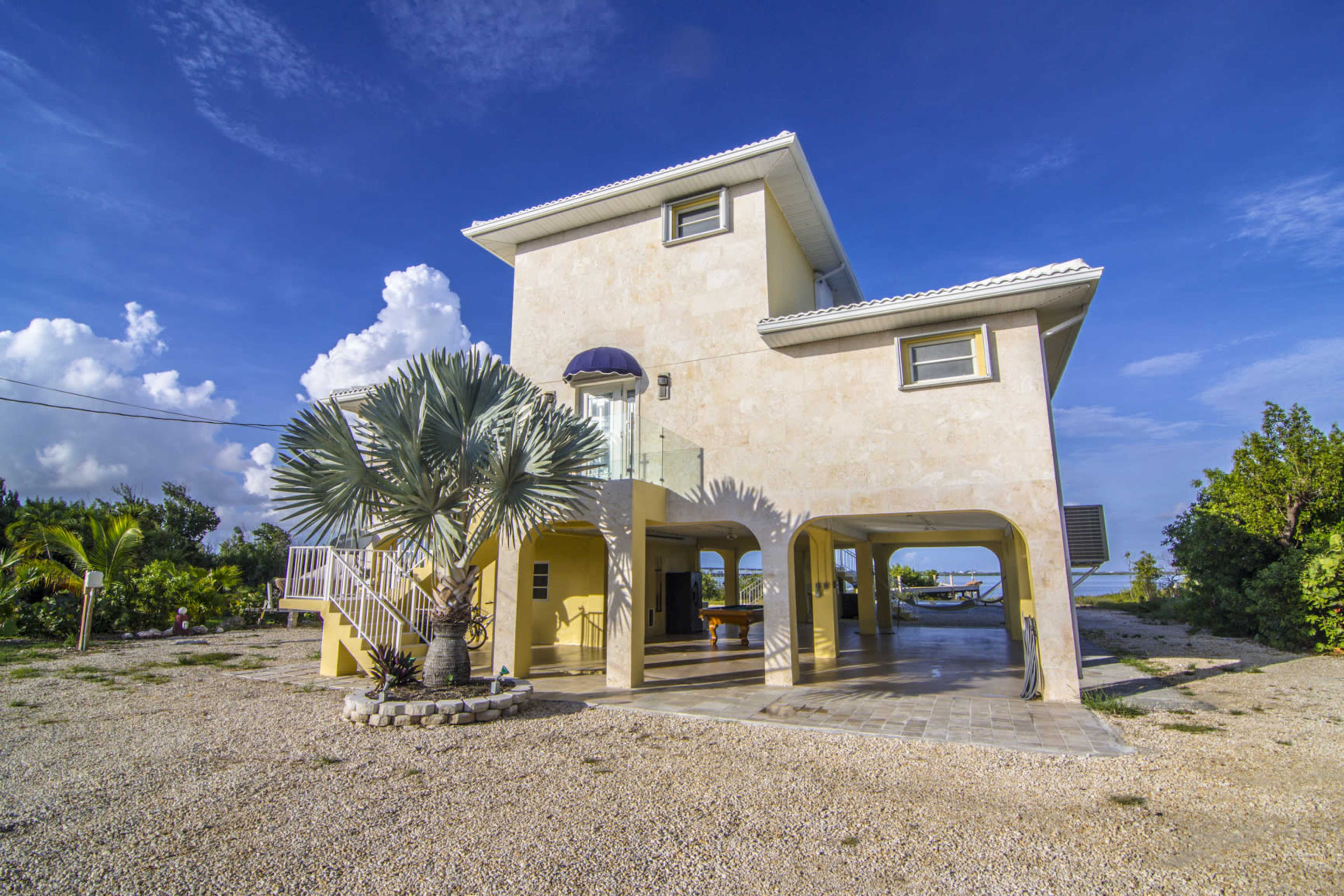 A two-story beige house with a balcony and open spaces underneath is surrounded by gravel, palm trees, and a clear blue sky.