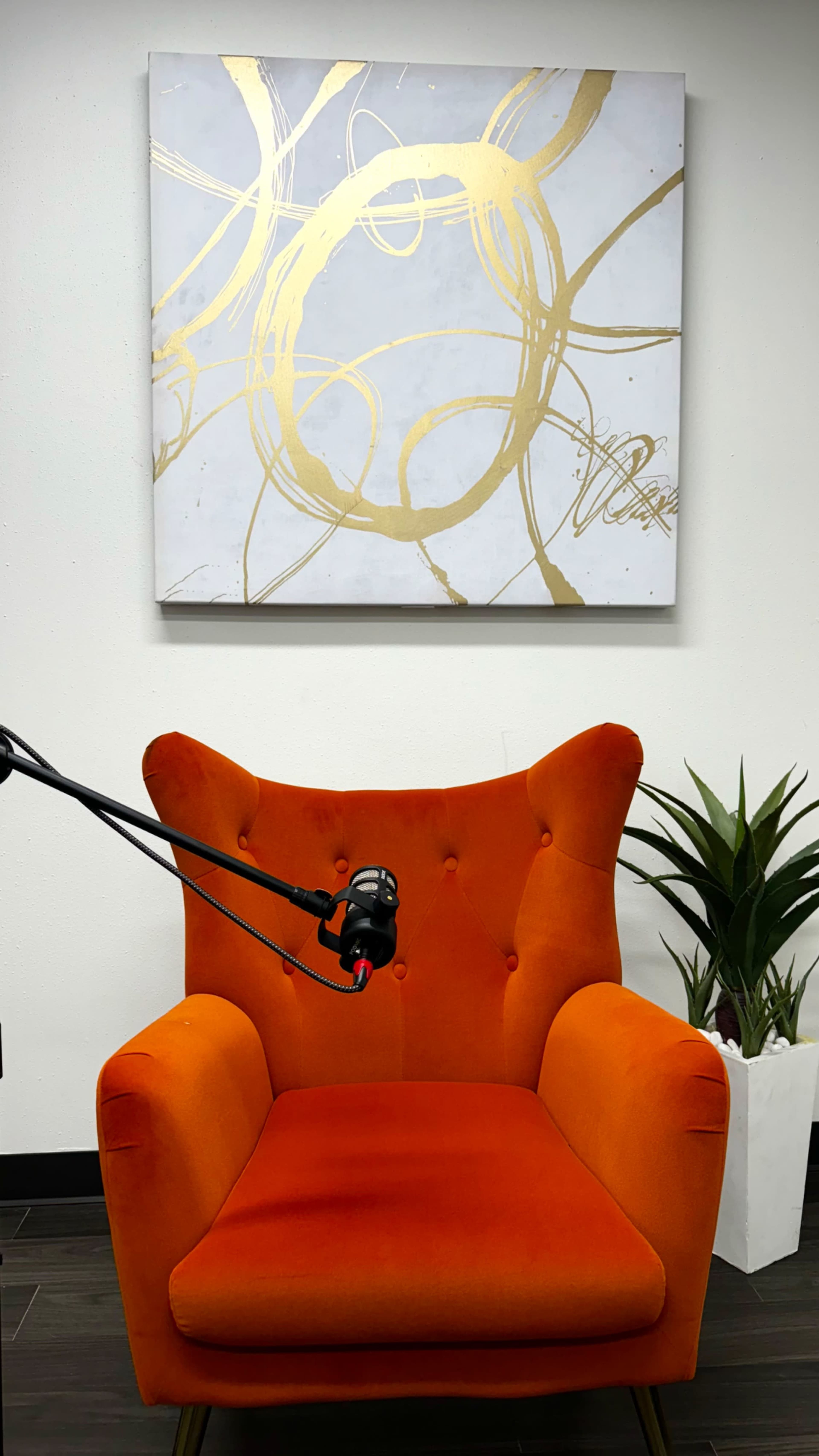 A bright orange chair with a microphone is positioned in front of a gold-accented painting and a green plant in a white pot.