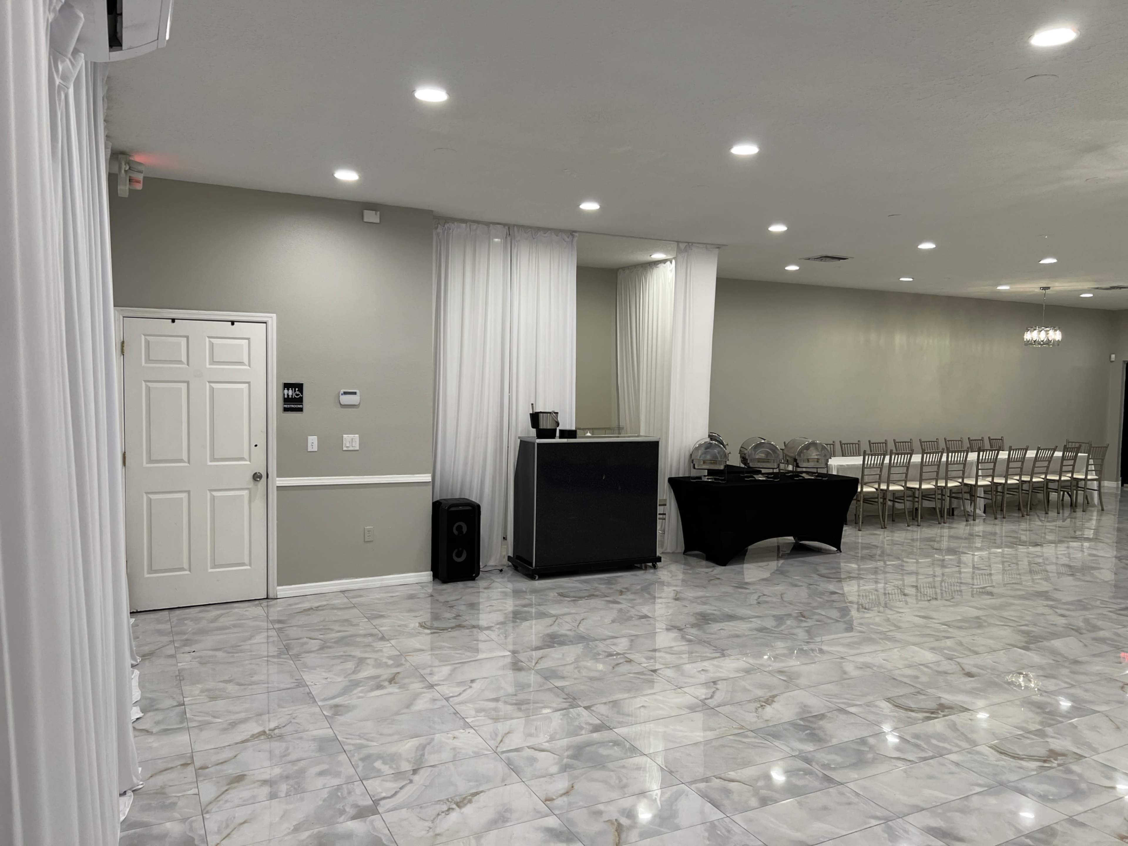 The image shows an event space featuring glossy marble floors, a seating area with arranged chairs, a refreshment station, and a door leading to another room.