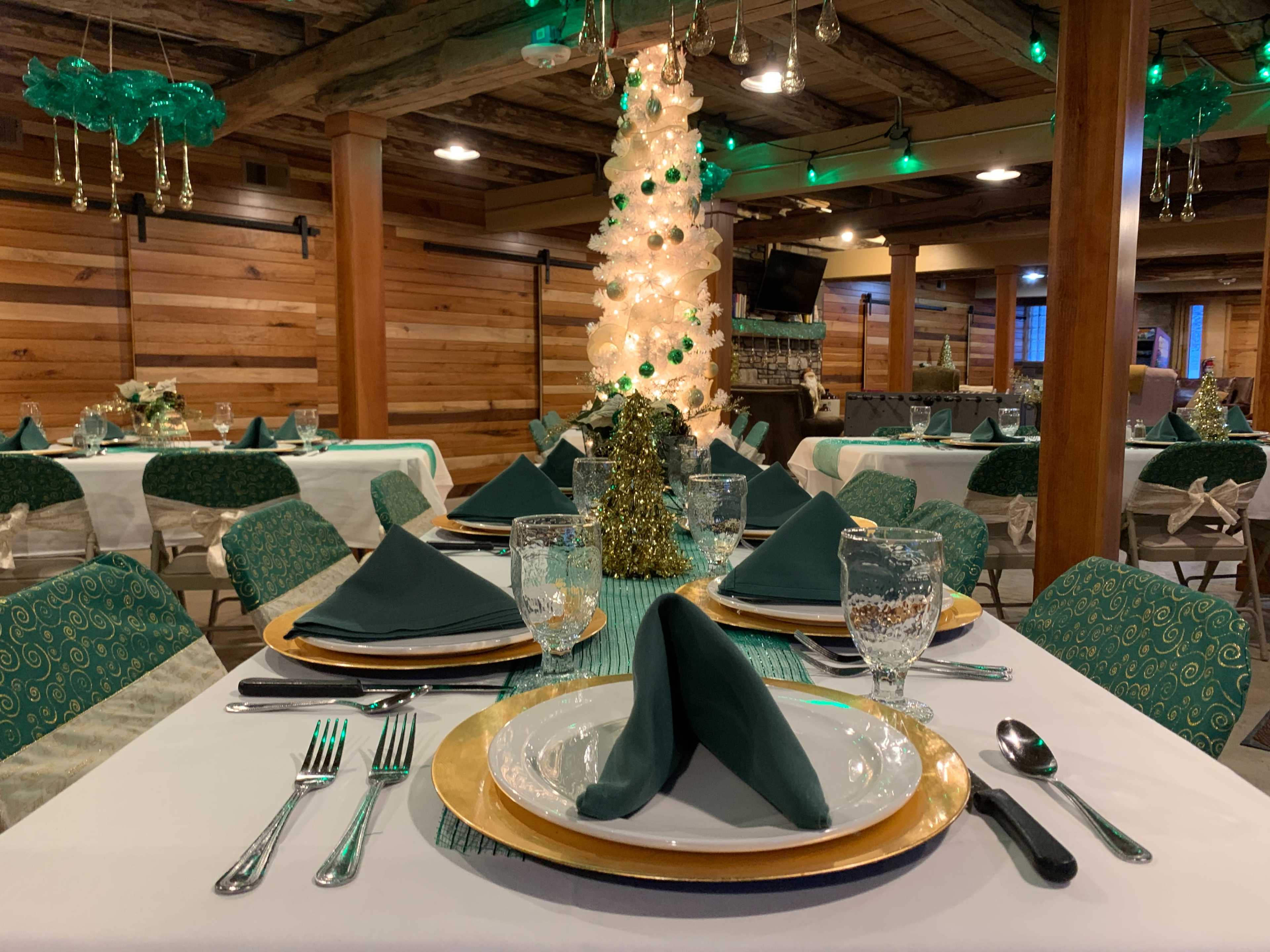 The image shows a dining setup in a rustic venue with tables arranged elegantly, featuring green napkins, gold-rimmed plates, and a decorated tree in the center.