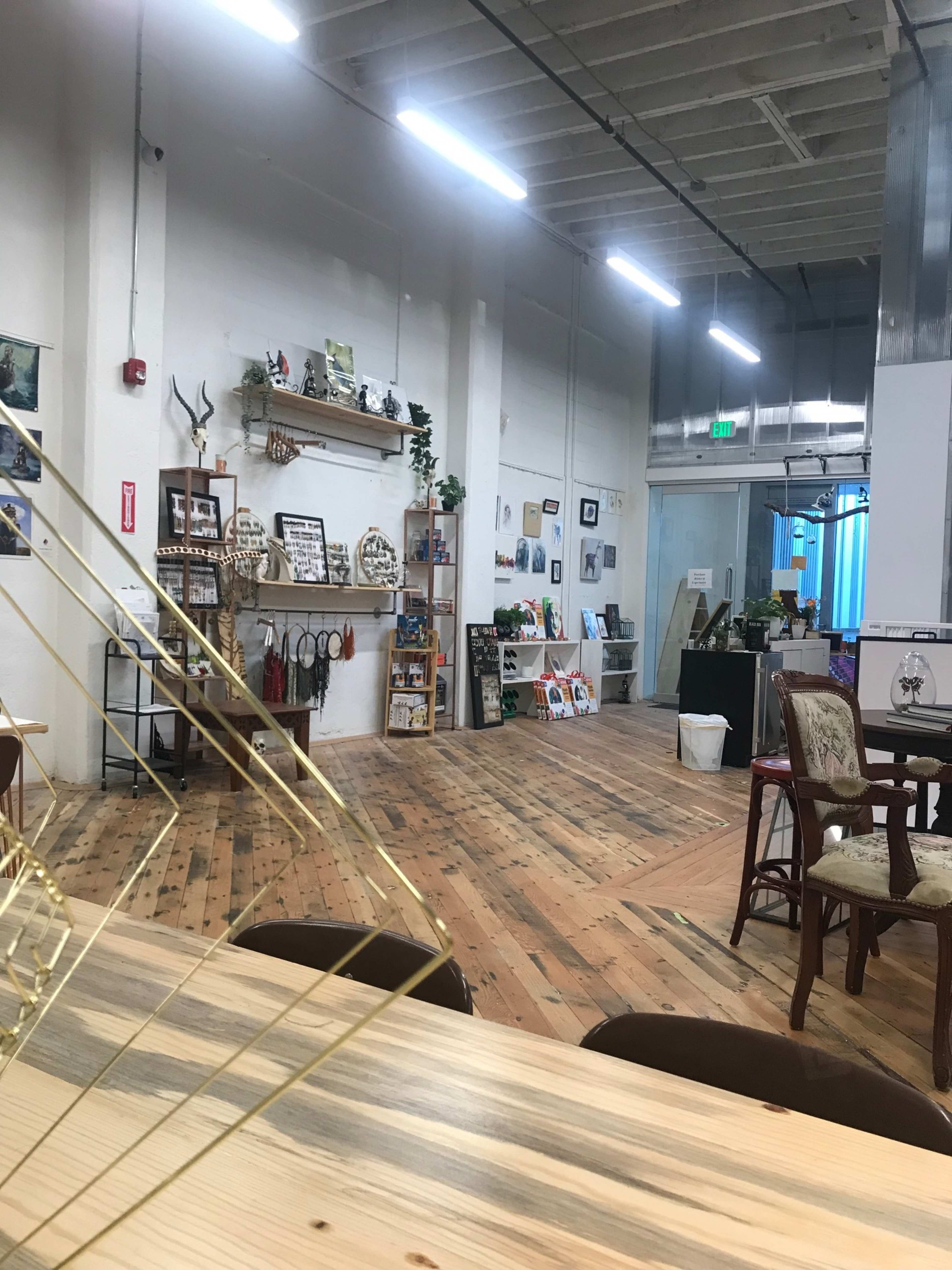 A spacious, brightly lit indoor area featuring wooden flooring, shelves with various decorative items, and a few tables and chairs arranged throughout.