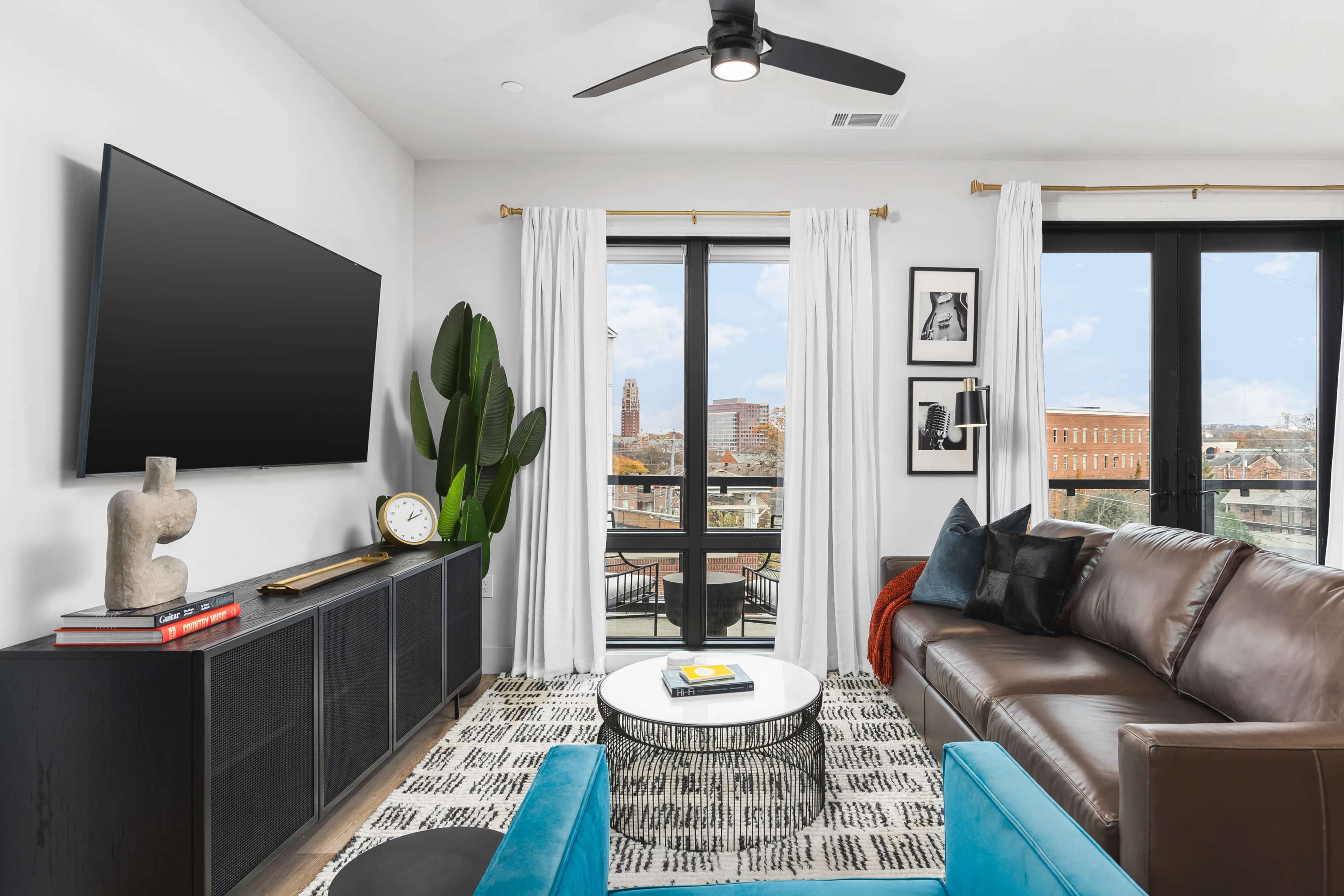 A modern living room with a large flat-screen TV mounted on the wall, a leather sofa, a round coffee table, and large windows offering a view of the city skyline.