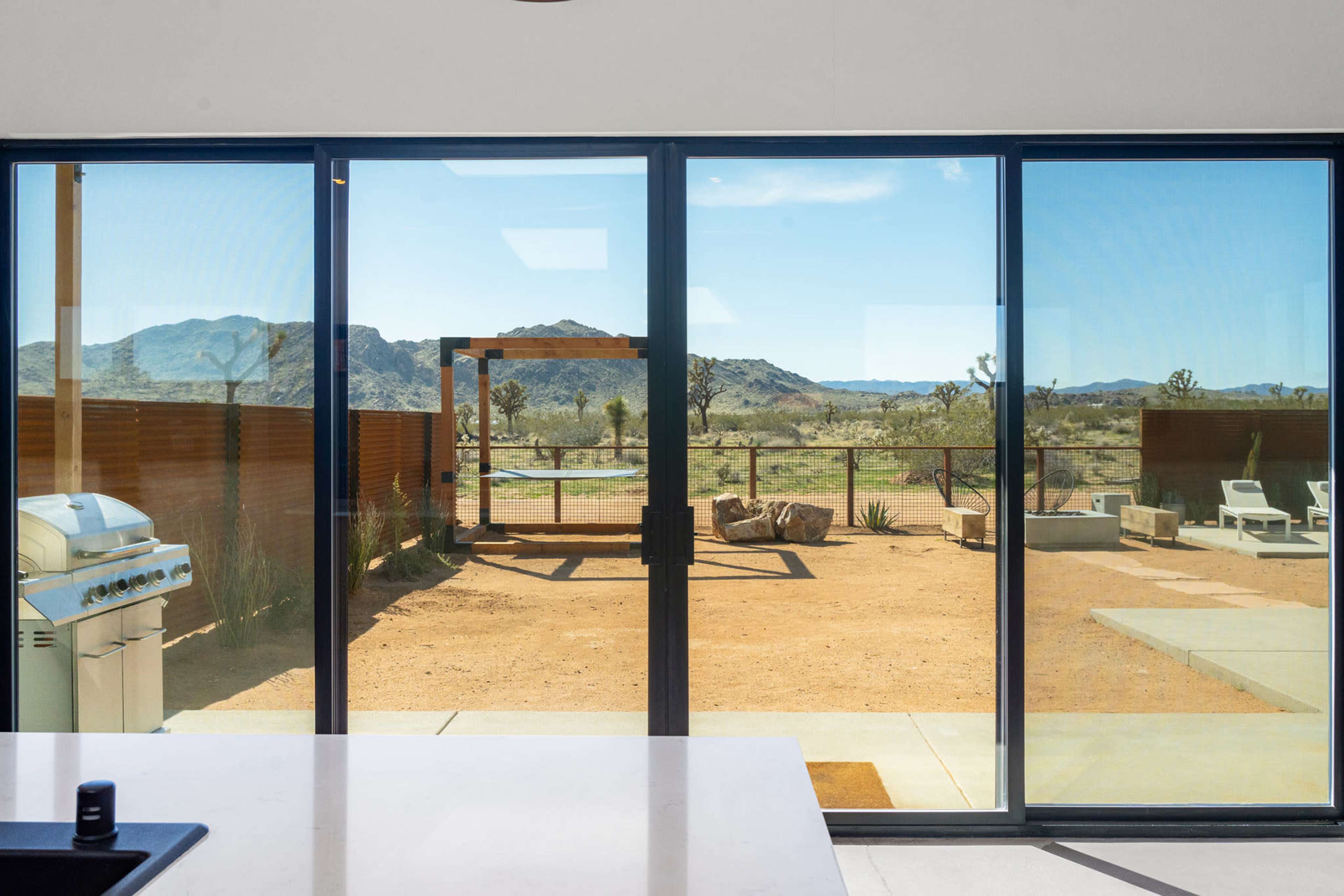 The image shows a large window view of a desert landscape featuring mountains, a patio area with lounge chairs, a barbecue grill, and a wooden structure in the foreground.