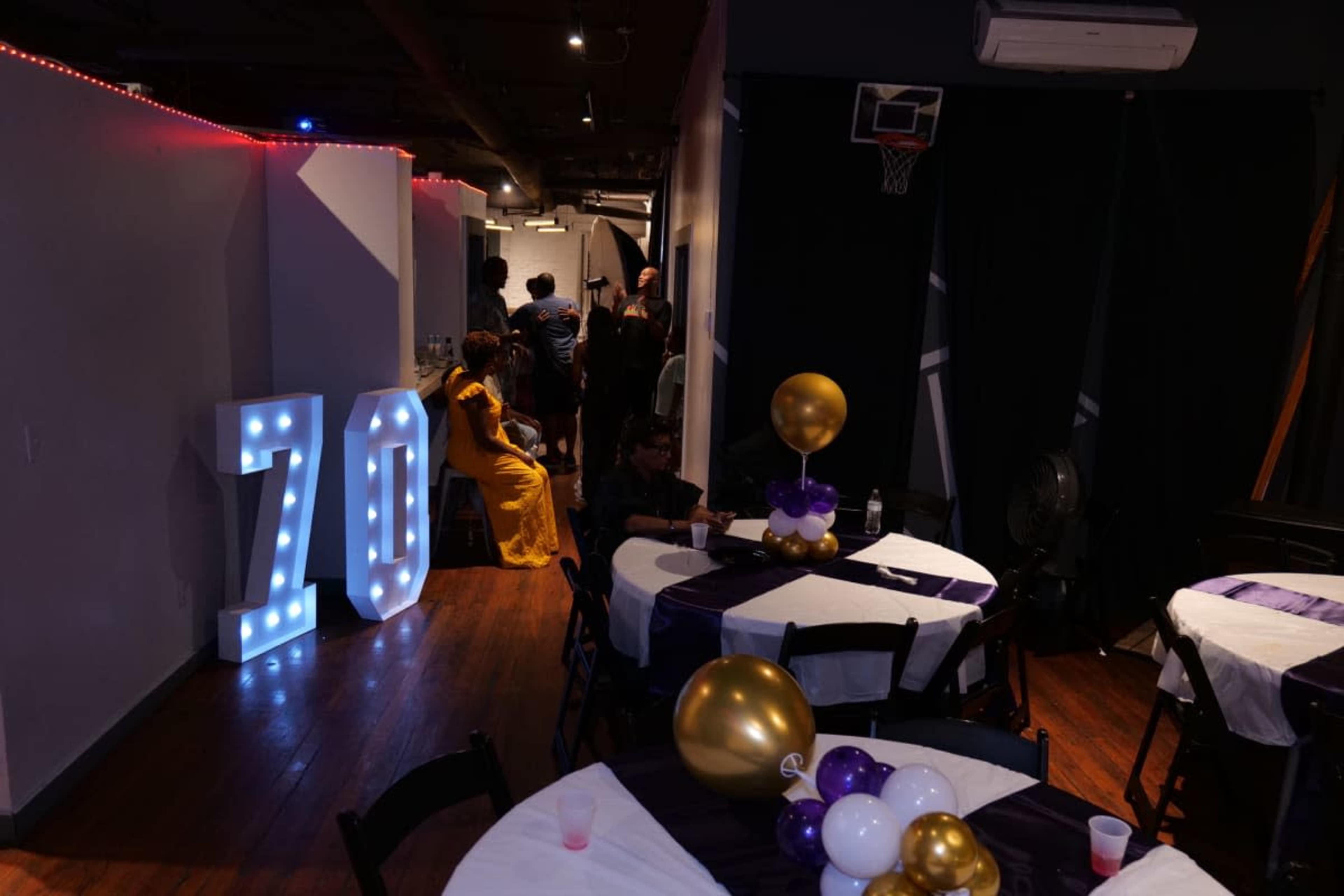 The image shows a decorated event space with tables set for a celebration, featuring large illuminated numbers "70" and balloons in gold and purple.