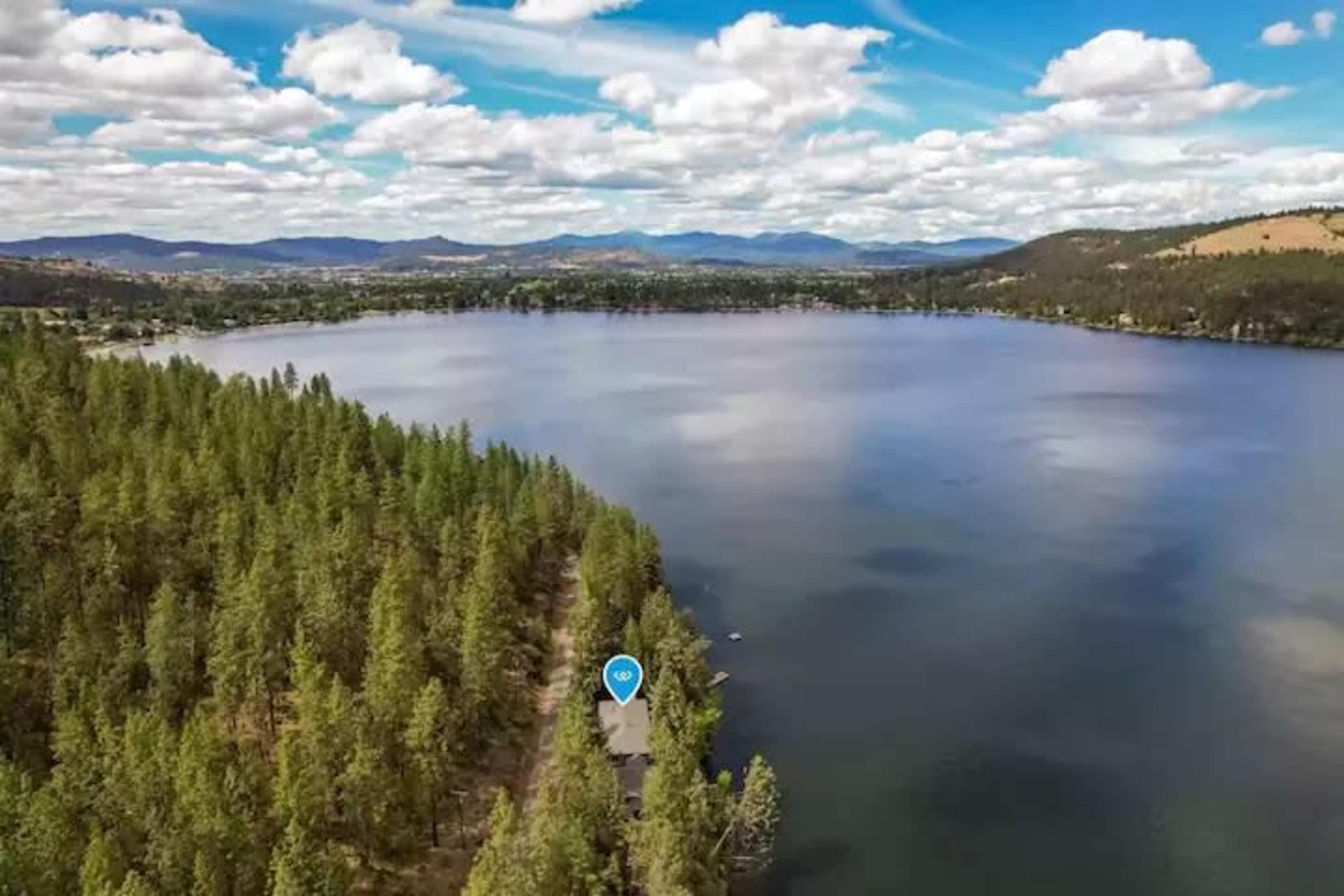 An aerial view shows a lake surrounded by wooded areas and distant mountains under a partly cloudy sky.