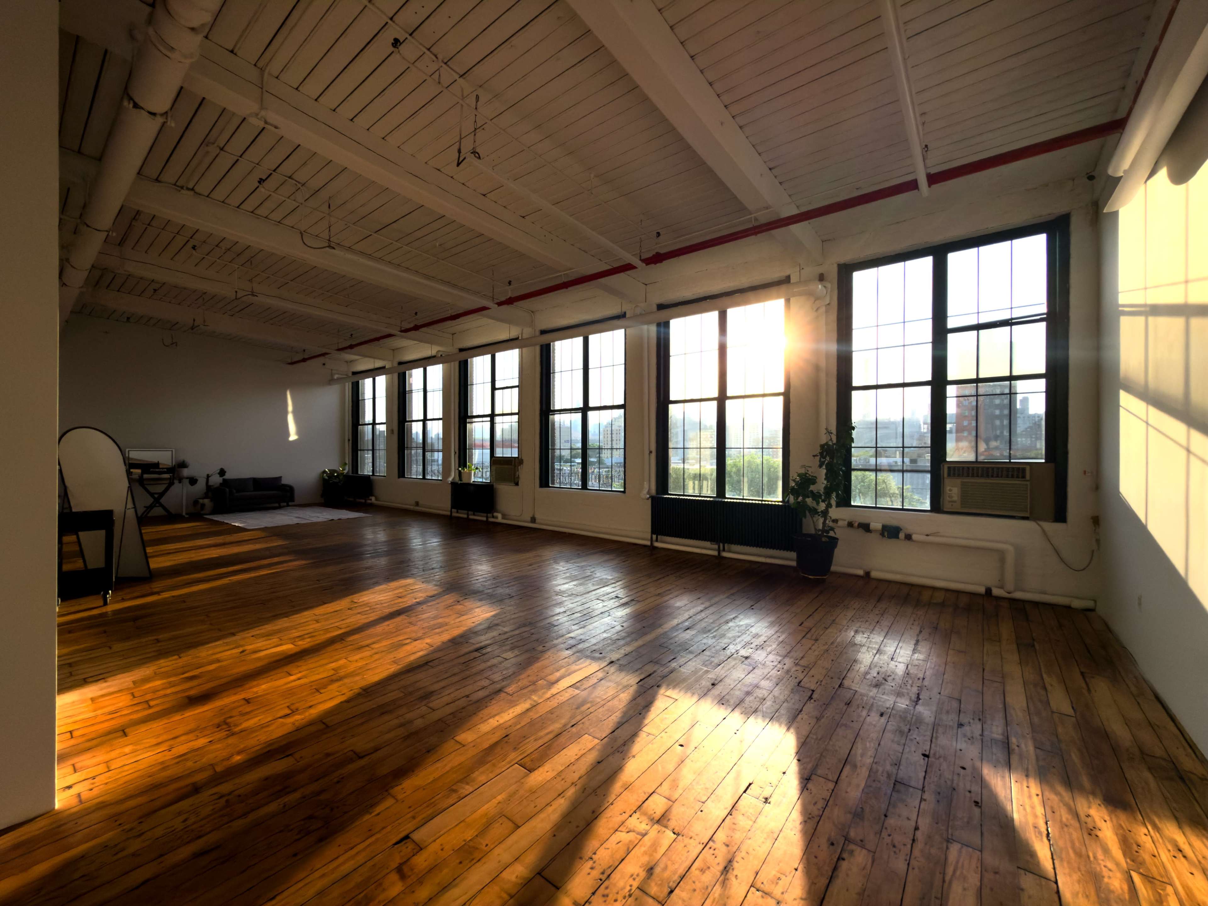 The image shows a spacious, sunlit room with wooden floors and large windows, showcasing an urban view.