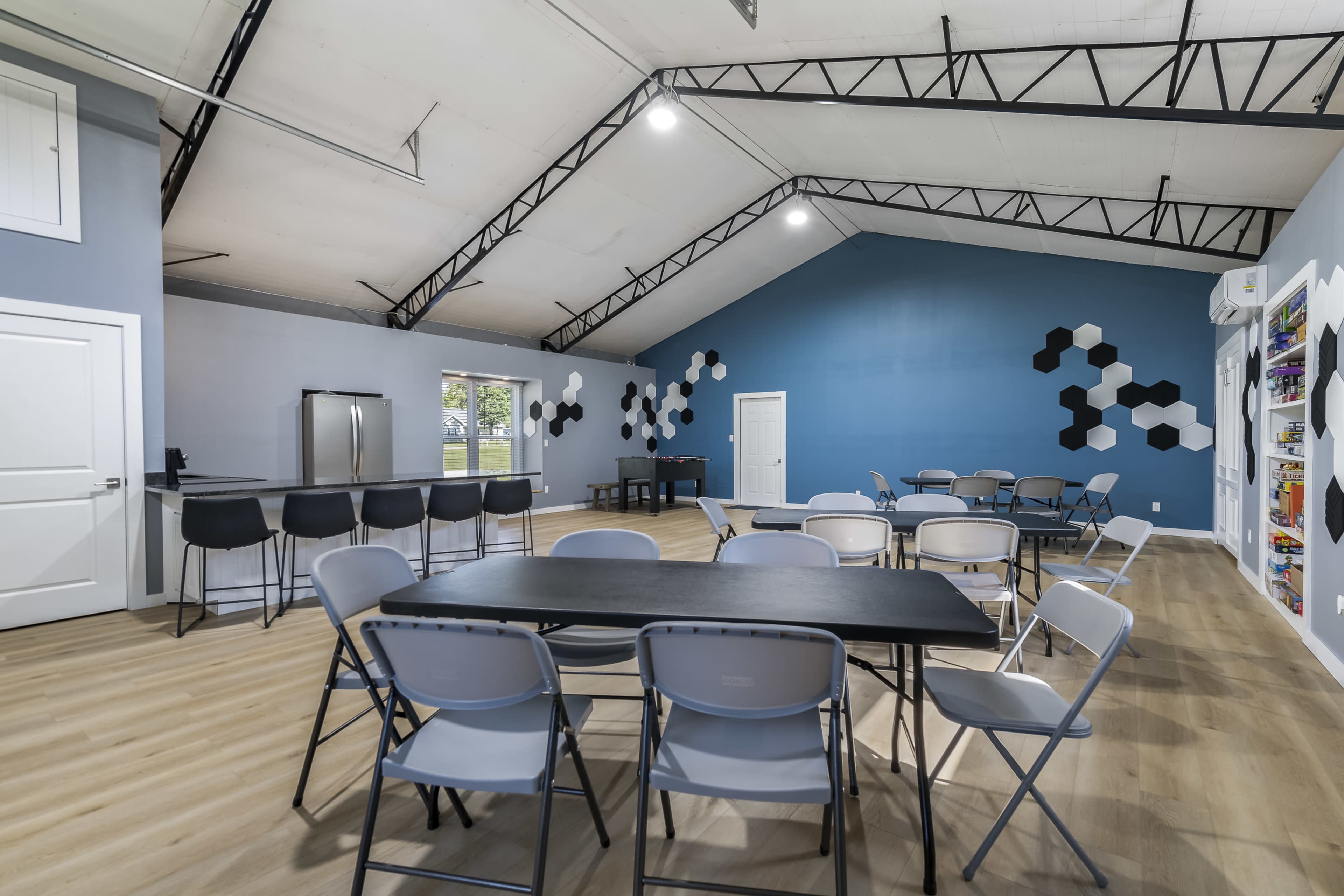 The image shows a spacious room with a high ceiling, featuring a set of tables and chairs, a kitchen area, and a blue accent wall decorated with geometric shapes.