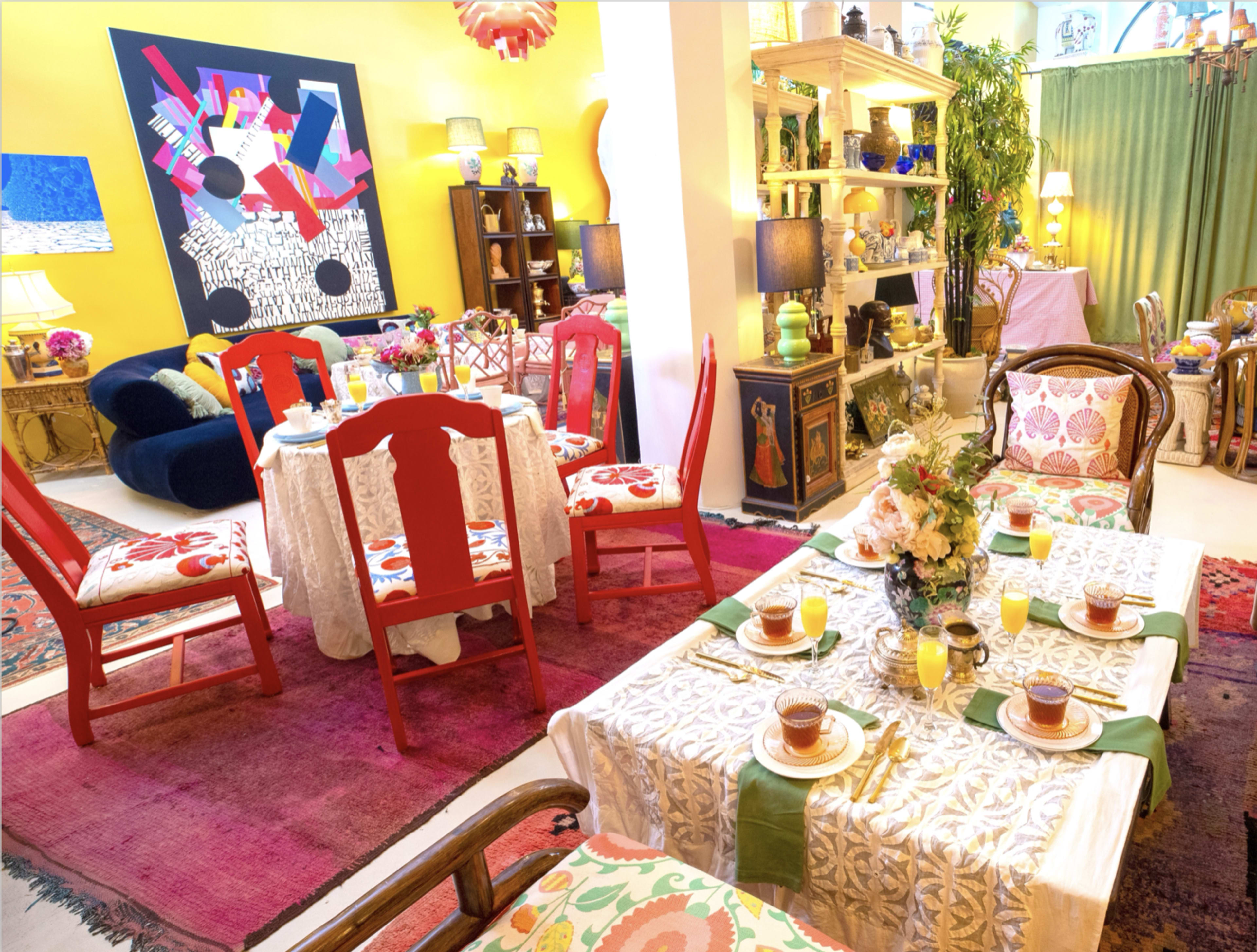 The image shows a brightly colored dining area with a table set for breakfast, surrounded by eclectic chairs and decorative shelves filled with various items.