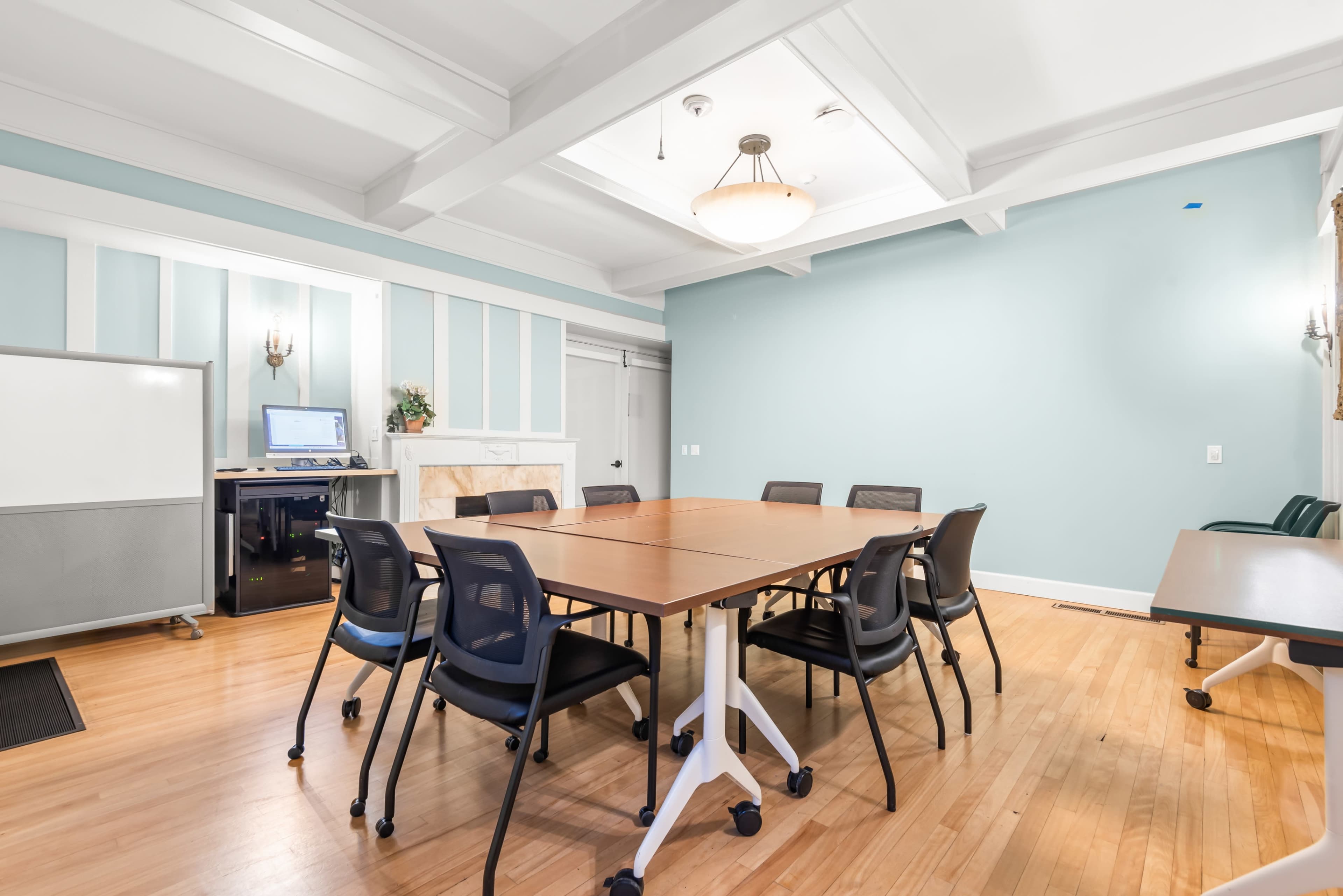 The image shows a well-lit meeting room with a large table surrounded by several black chairs, a whiteboard, and a computer station in the corner.