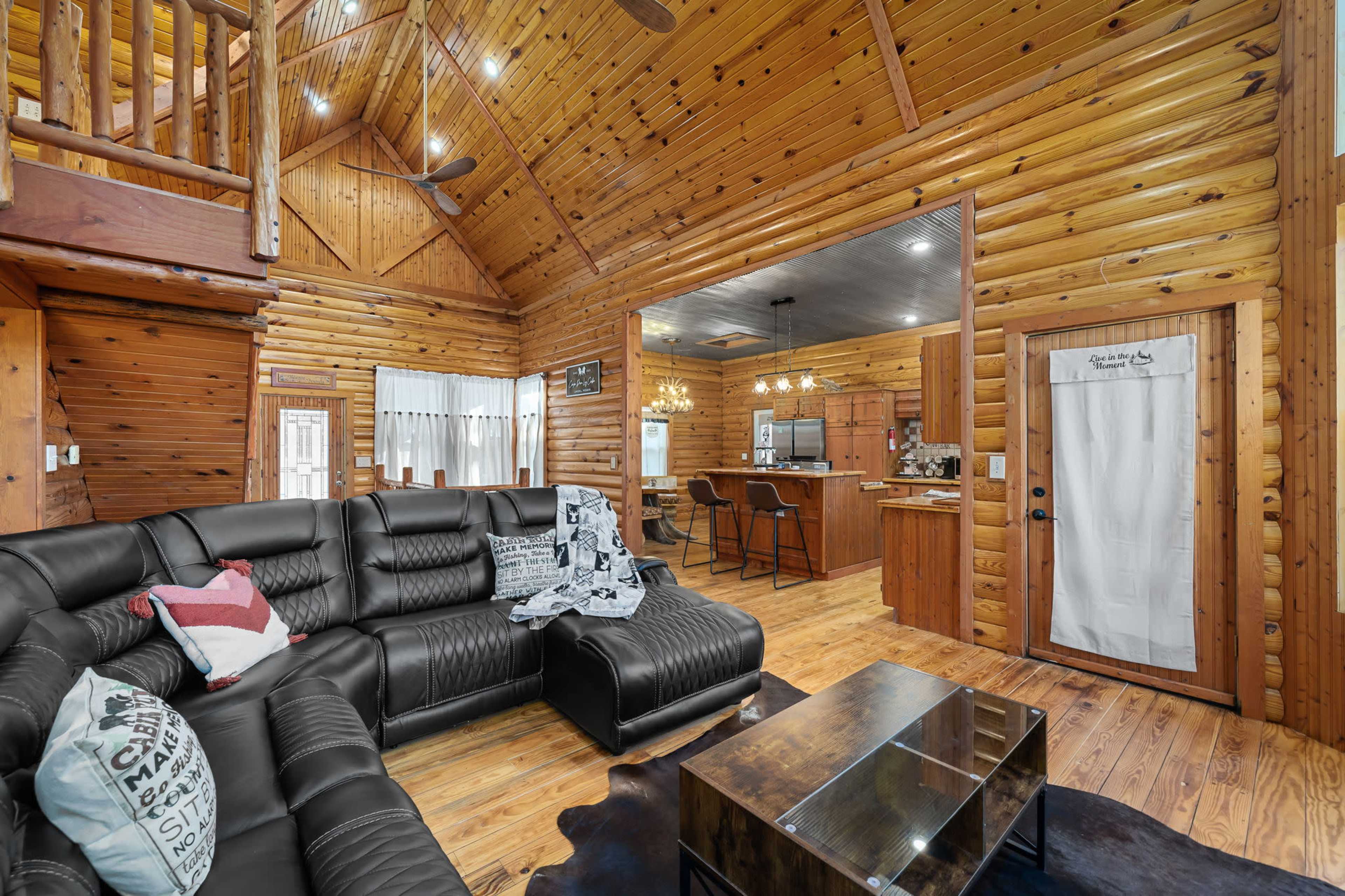 A spacious, wooden interior of a cabin with a large black sectional sofa, a glass coffee table, and an open kitchen area in the background.