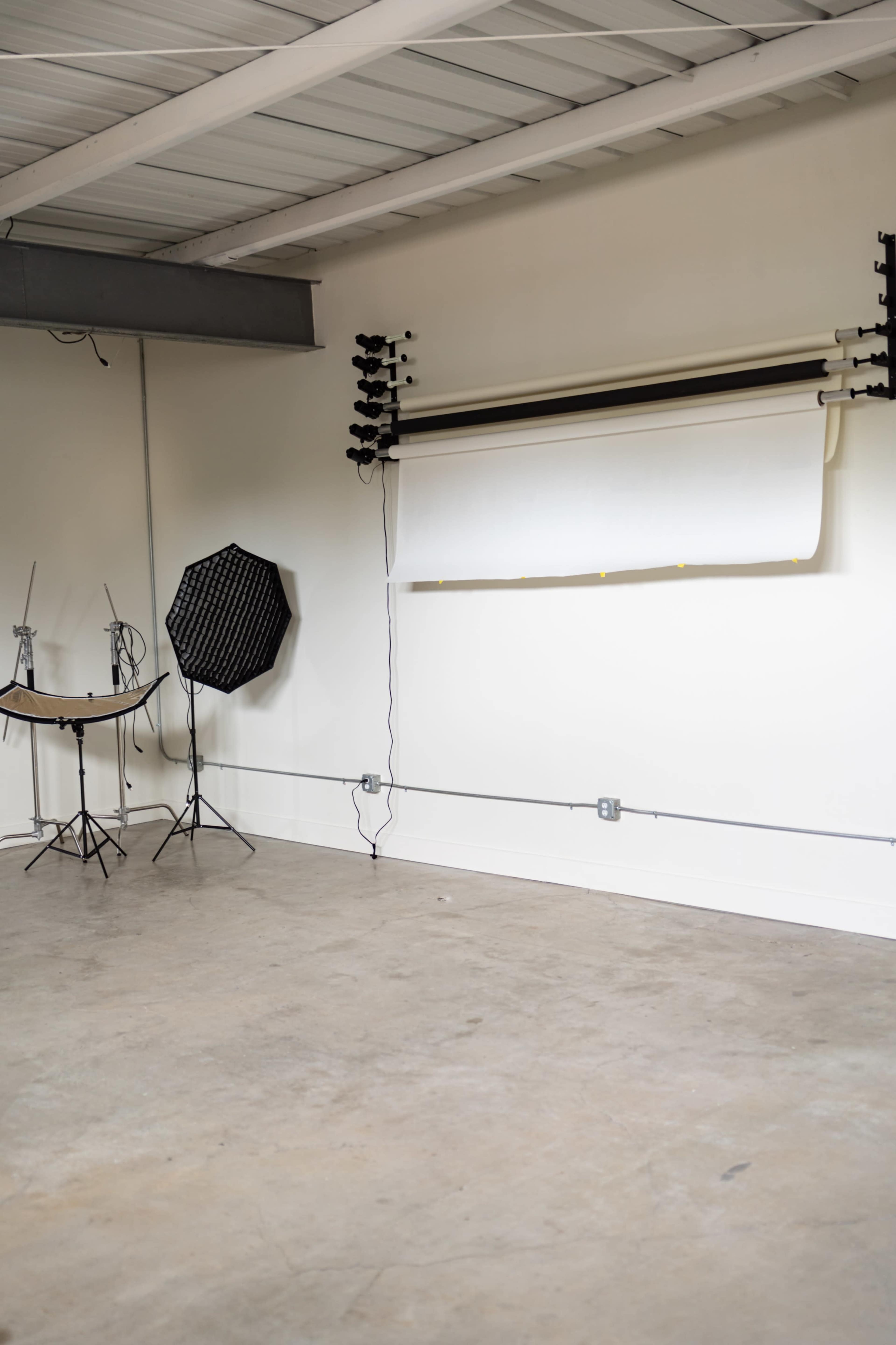 The image shows a sparse studio space featuring a backdrop setup with a rolled white paper, a folding table, and lighting equipment against a plain white wall.