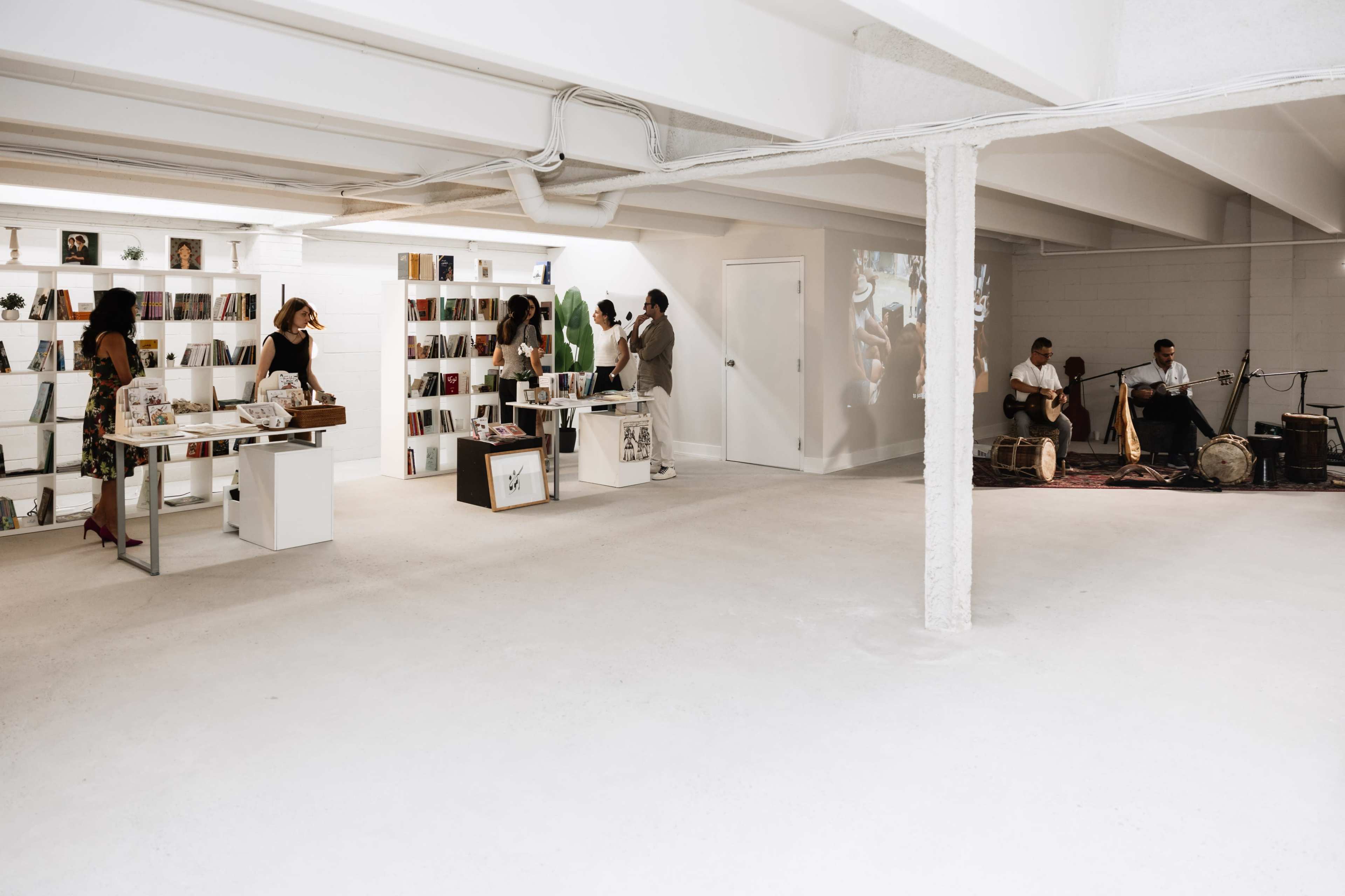 The image shows a spacious white-walled basement gallery featuring bookshelves, visitors browsing, and a live music performance taking place in one area.