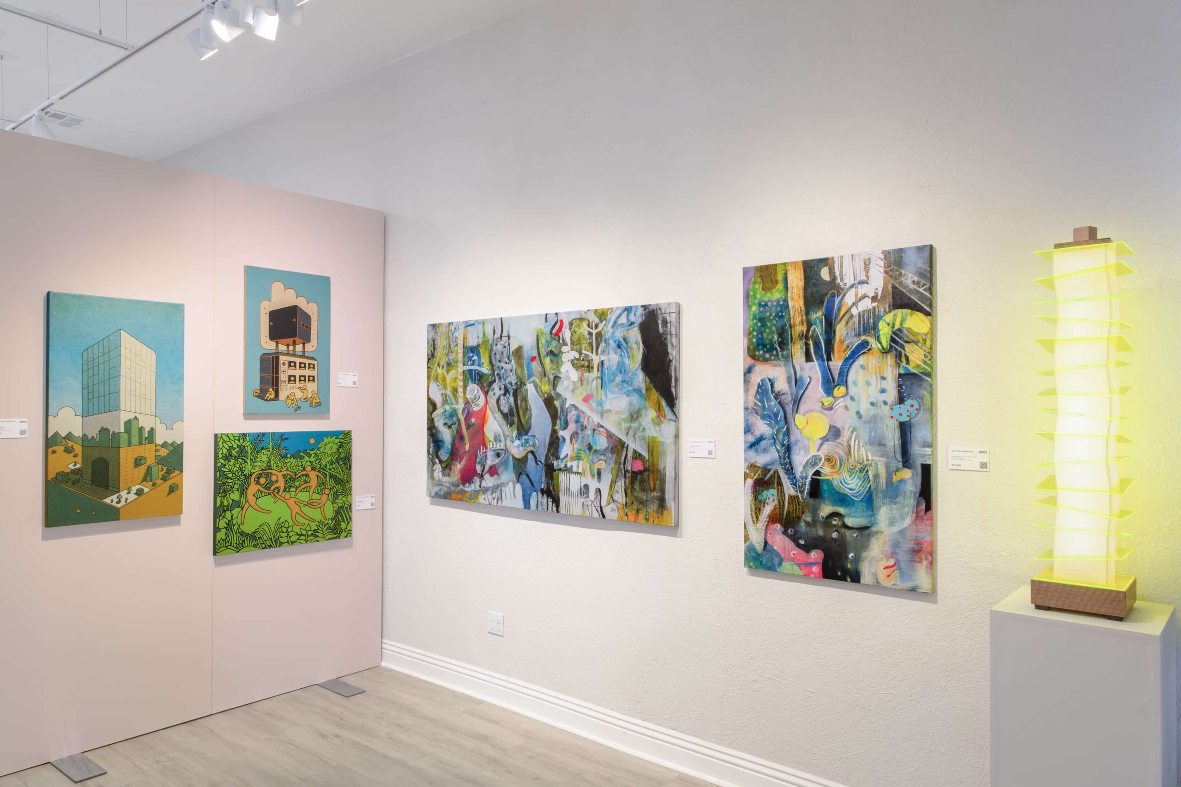 The image shows an art gallery featuring colorful paintings on pink and white walls, along with a unique illuminated lamp on a pedestal.