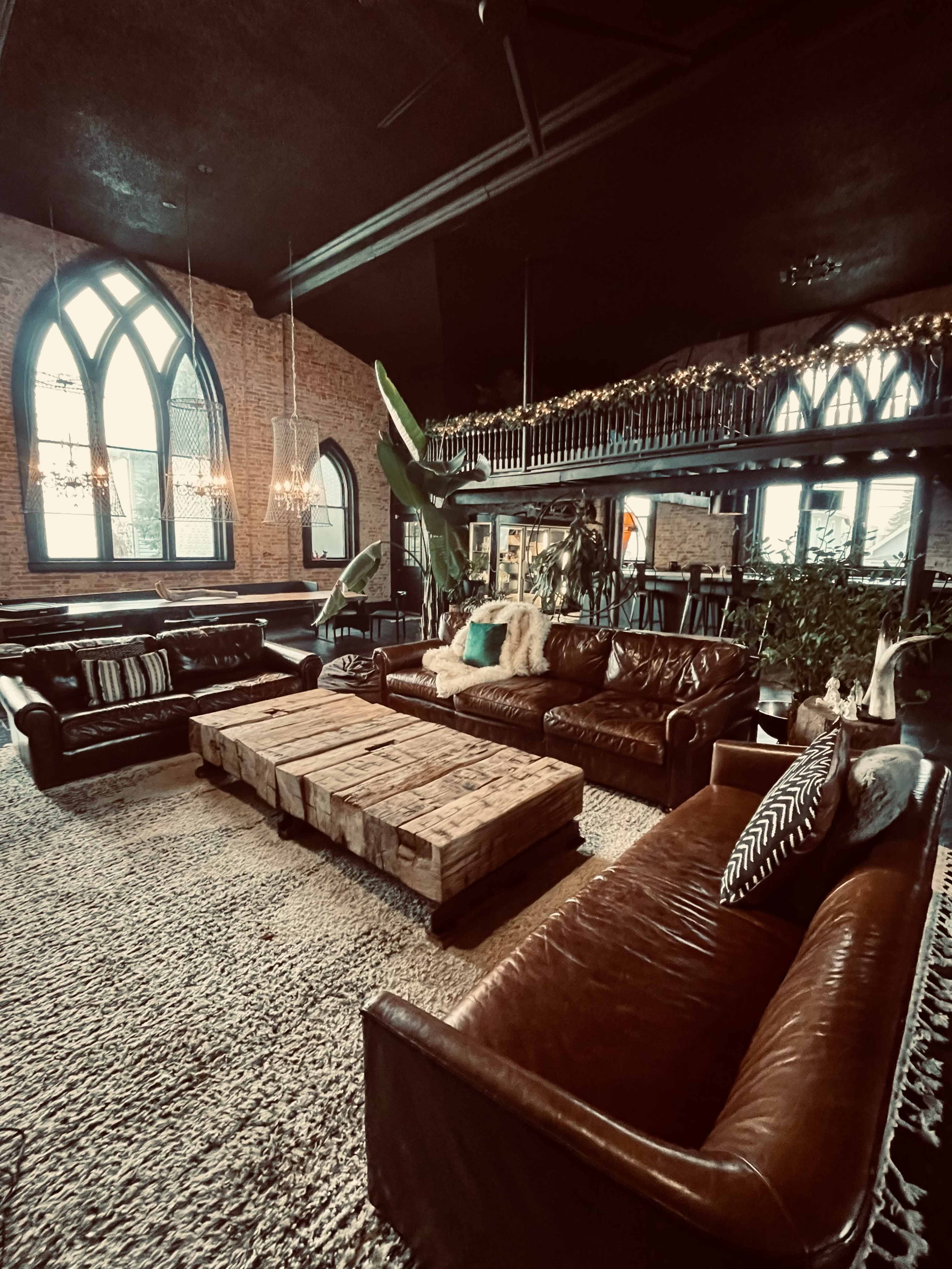 A cozy lounge area with brown leather sofas, a large wooden coffee table made from pallets, and a lush plant, set against a backdrop of brick walls and arched windows.
