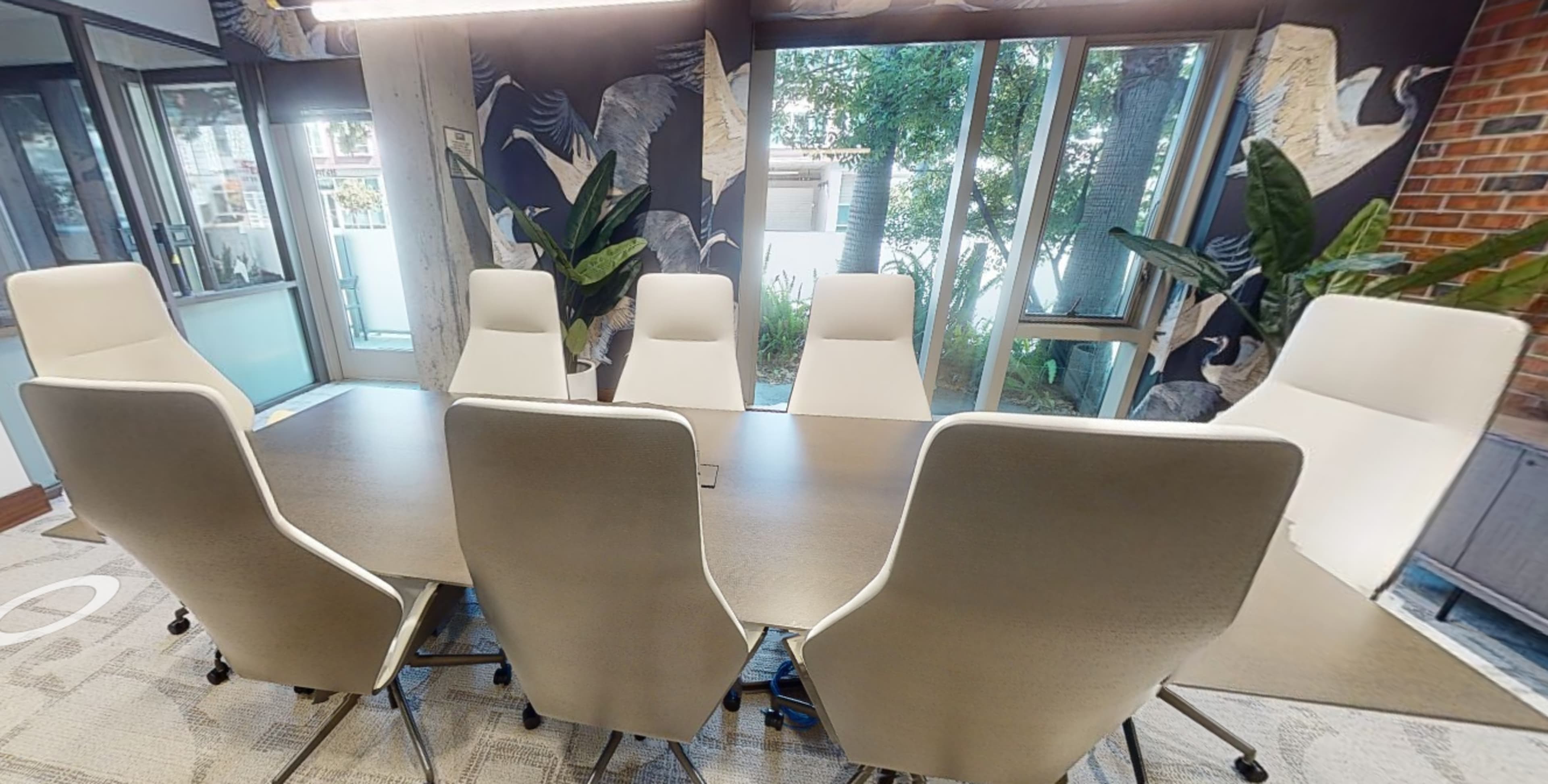A modern conference room features a large table surrounded by high-backed chairs and large windows with plants in the background.