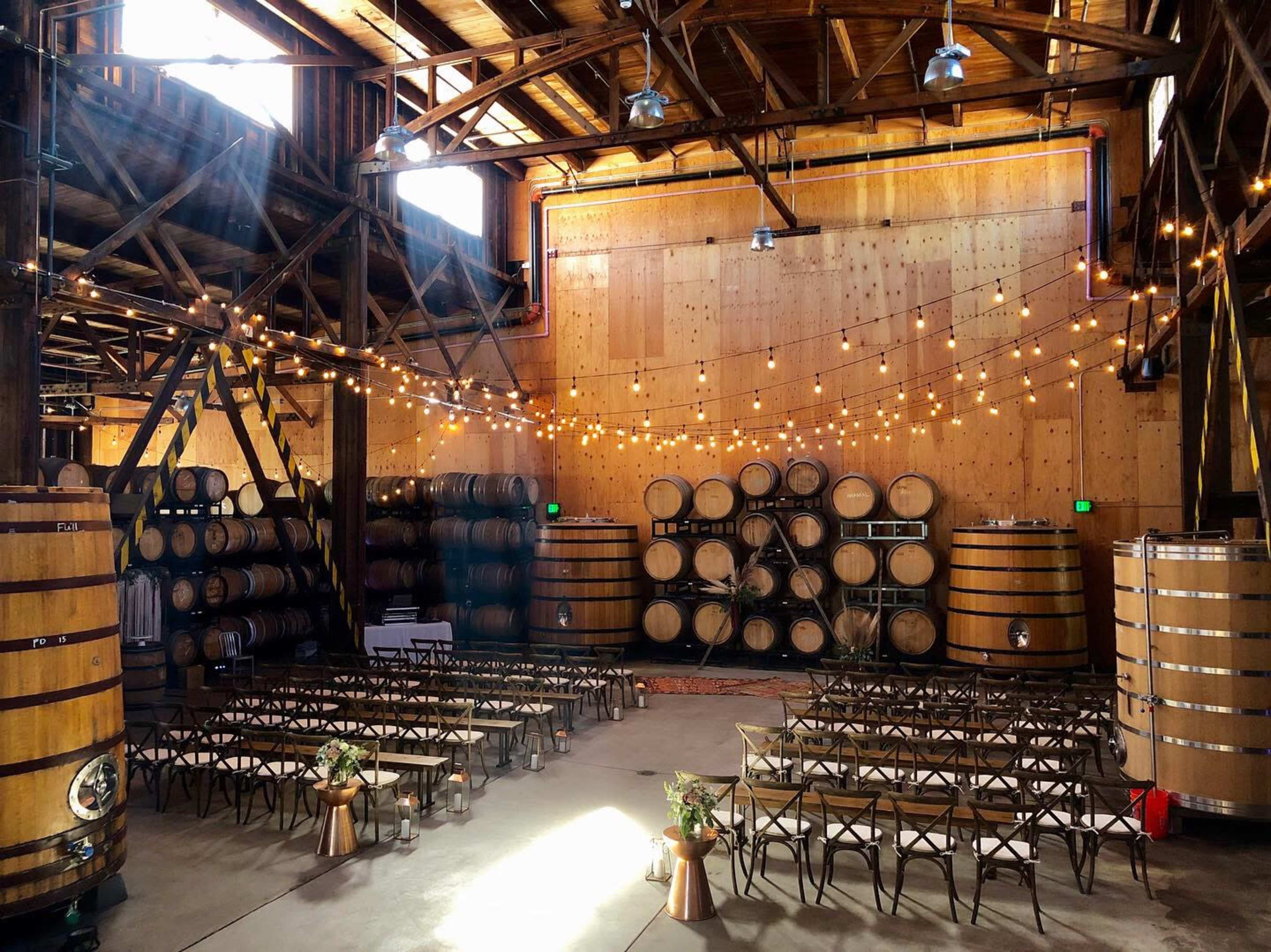 The image shows a spacious event venue featuring rows of seating in front of large wine barrels and rustic wooden walls, illuminated by string lights.