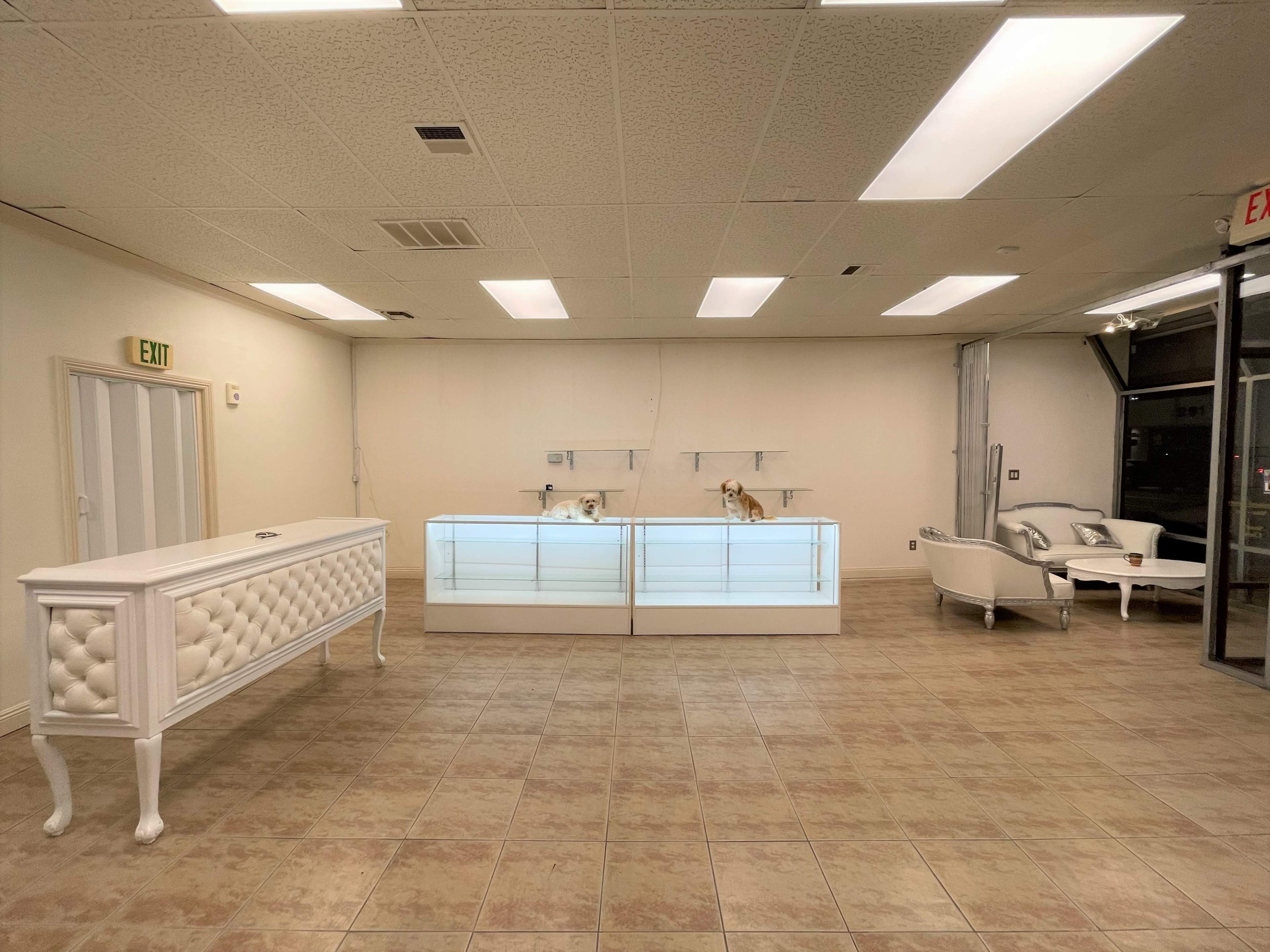 A sparsely furnished retail space featuring a white display counter with glass tops, two glass display cases, and a seating area with a white couch and a coffee table.