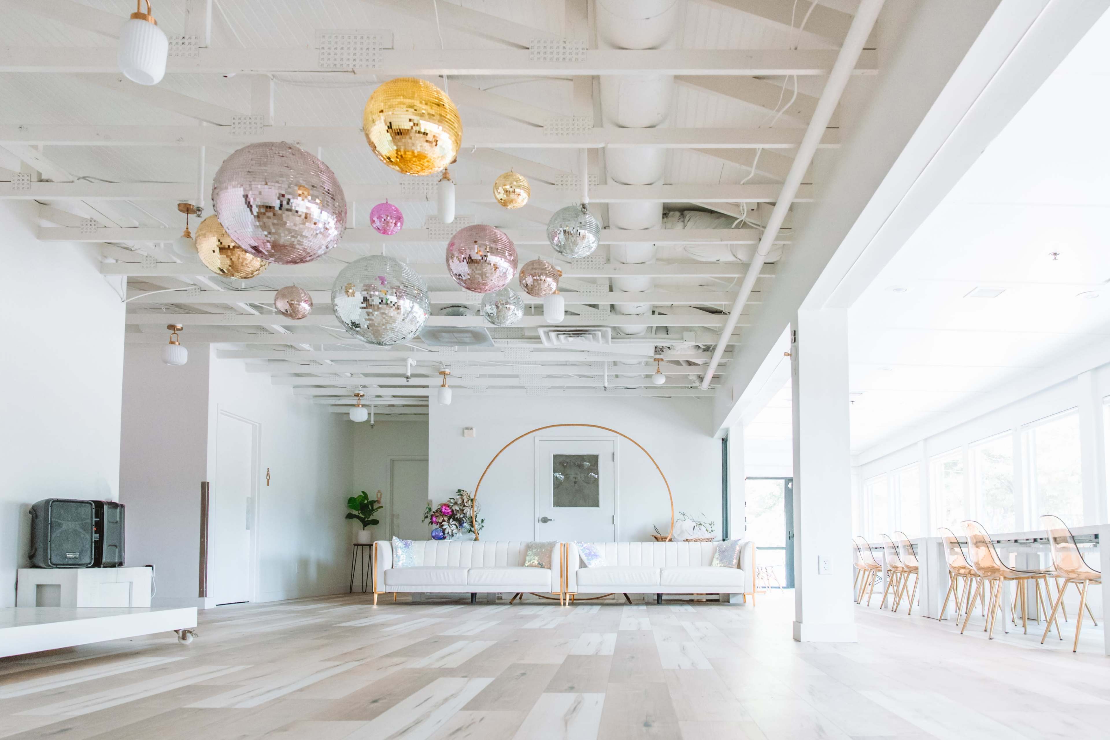 A bright, open space featuring a seating area with a sofa, decorative plants, and large hanging disco balls above hardwood flooring.