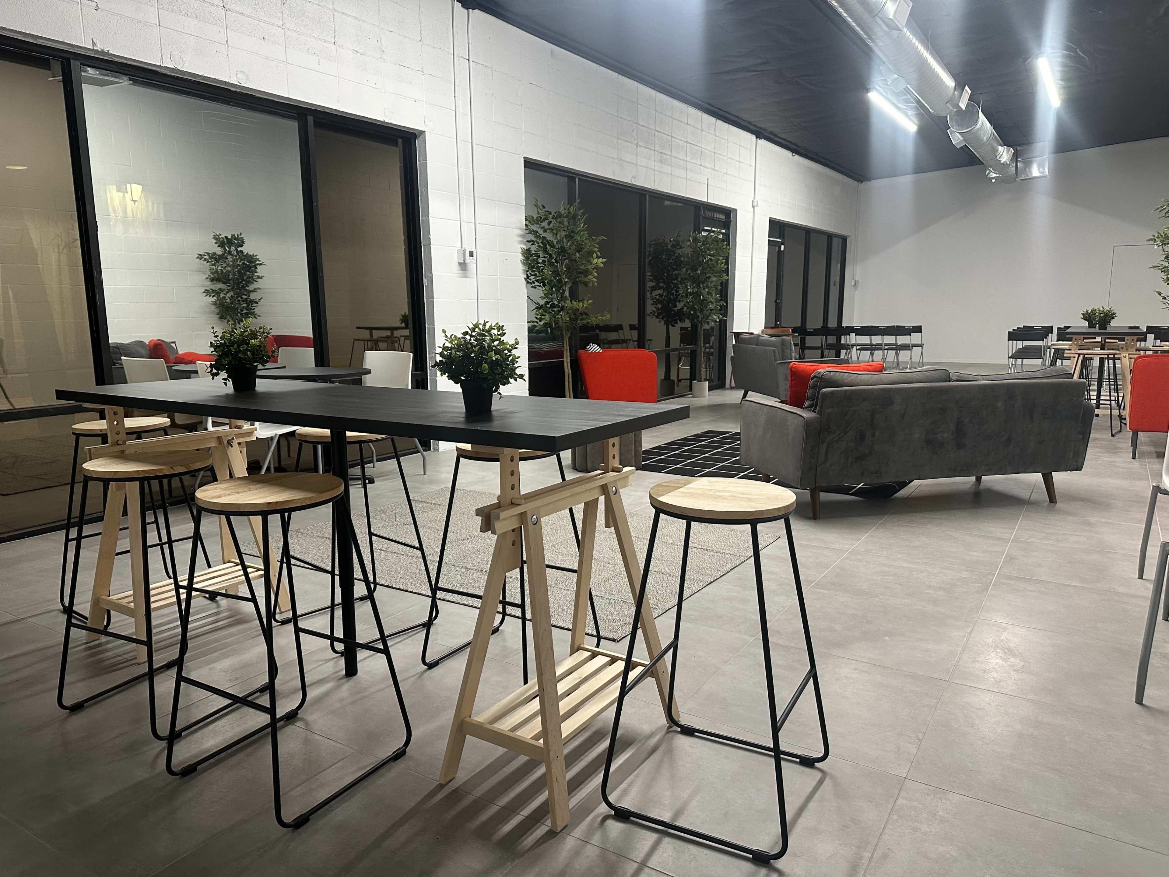 A modern, open space with high tables and stools, a sofa area, and potted plants arranged throughout.