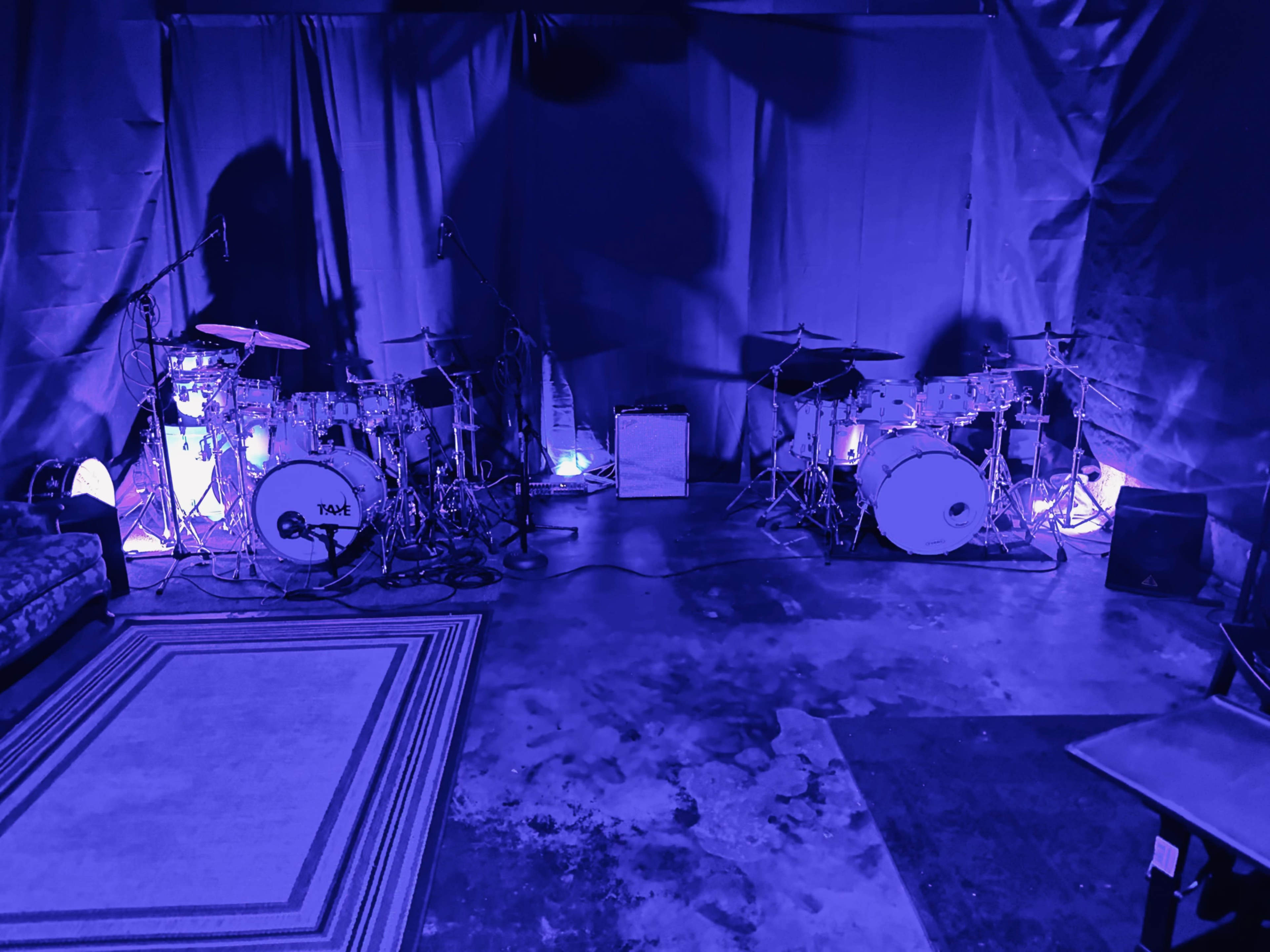 A music rehearsal space features two drum sets illuminated by purple lights, with microphones and an amplifier positioned nearby.