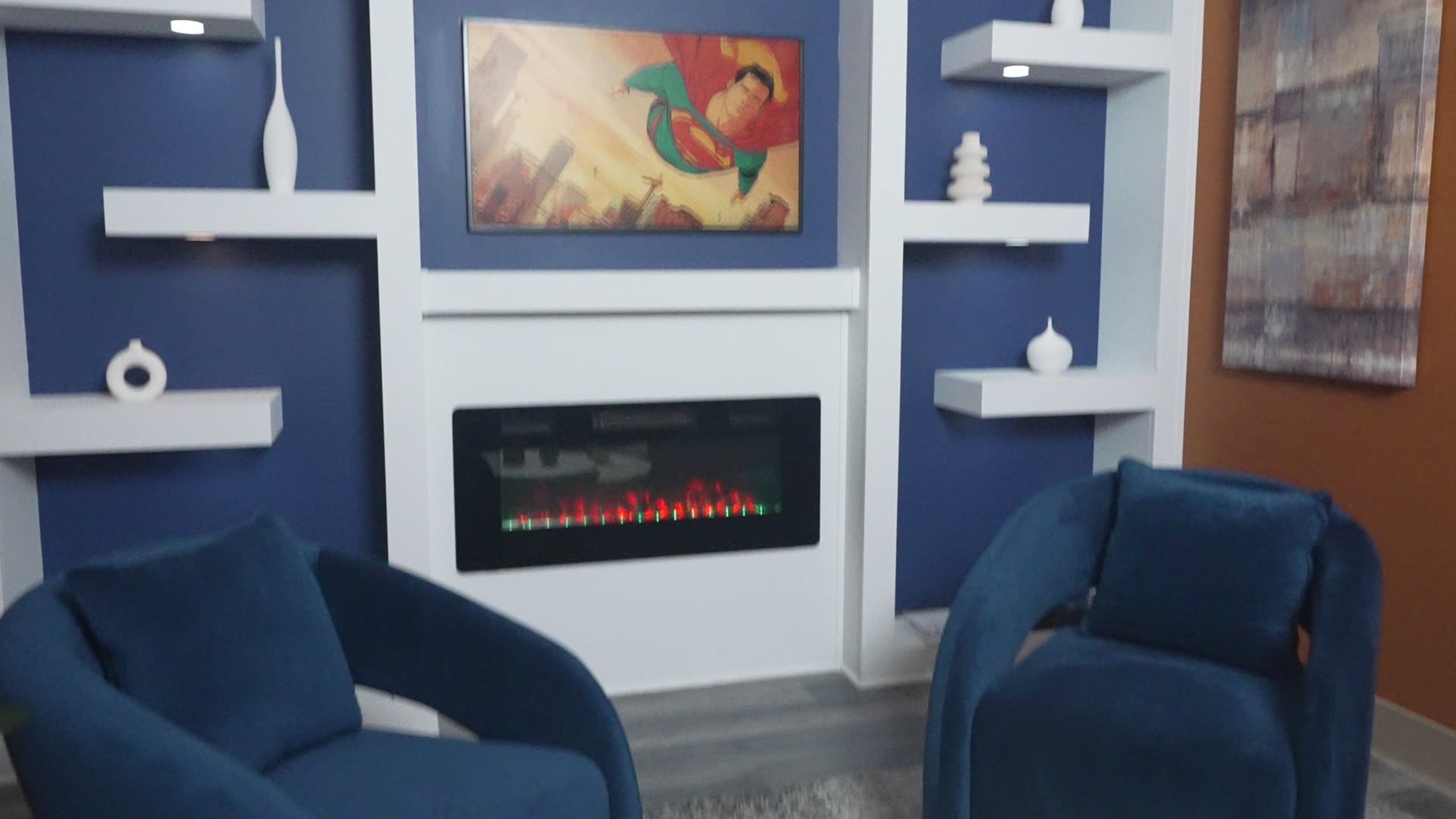 The image shows a modern living space featuring two blue chairs, a wall-mounted electric fireplace, and a Superman-themed artwork above it, with white shelves and decorative items on the blue wall.