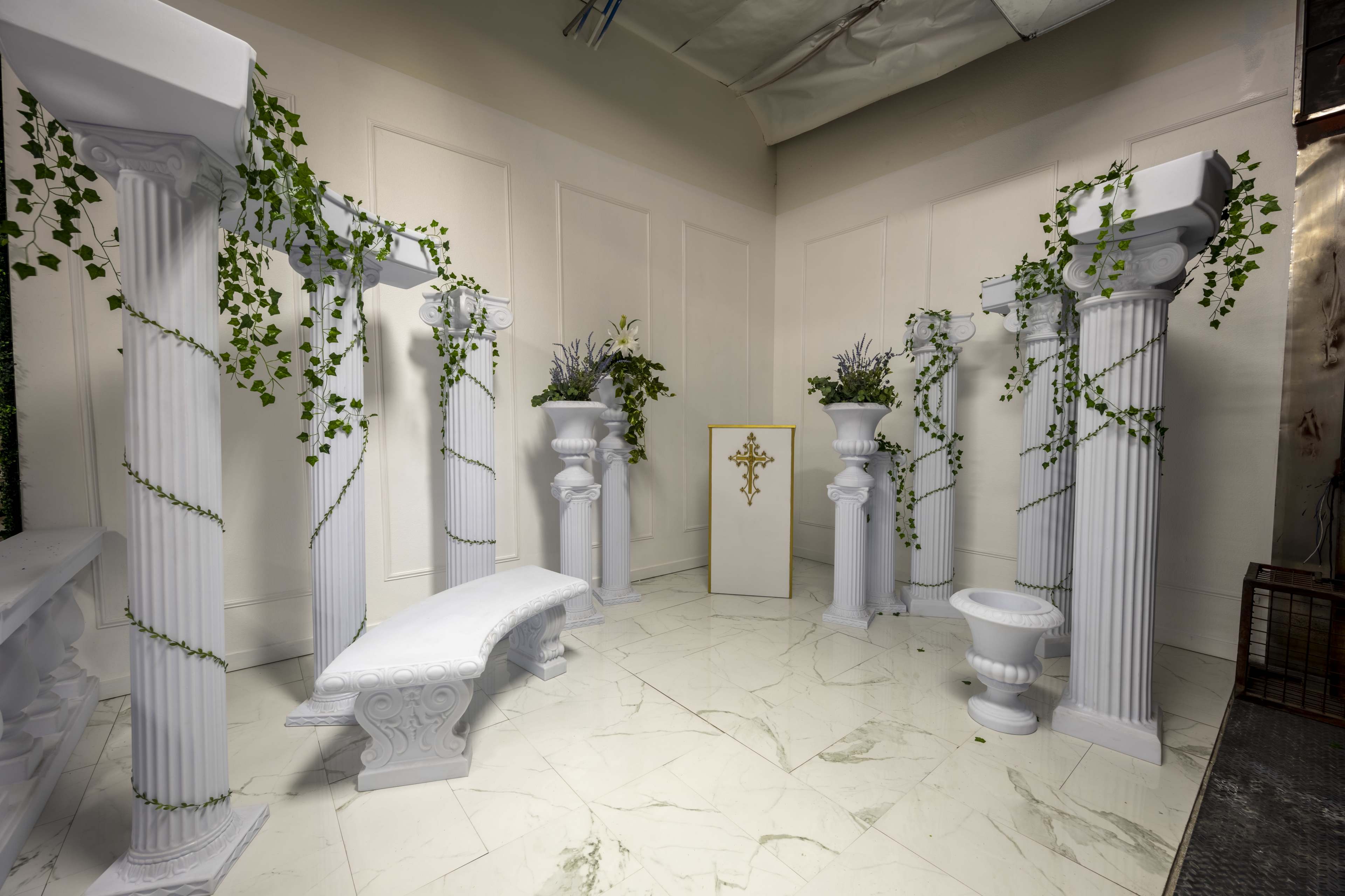 A white, columned space decorated with green vines, featuring a central decorative structure and marble flooring.
