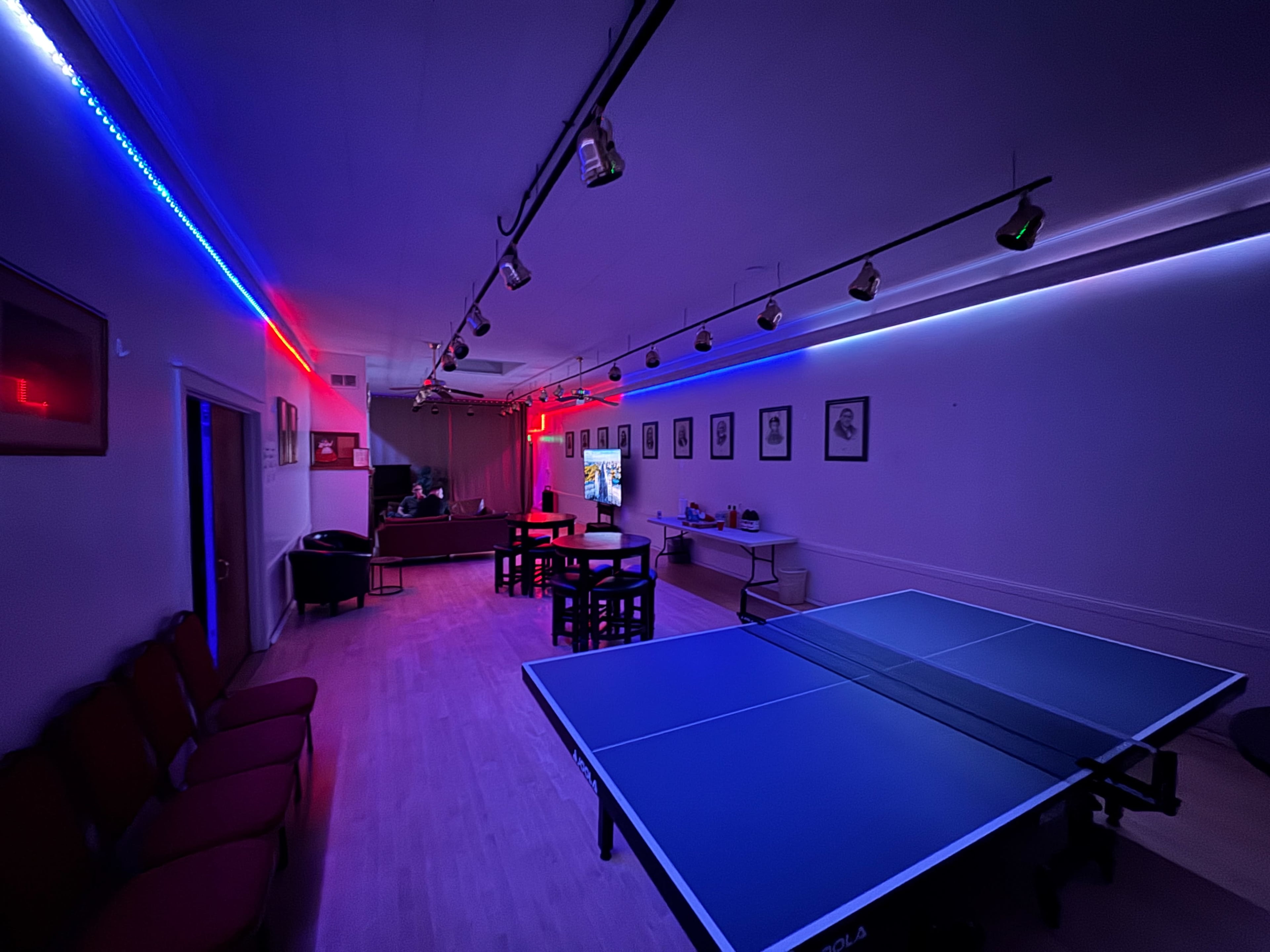 The image shows a dimly lit room with a blue ping pong table in the foreground and a bar area in the background, illuminated by colored lights.