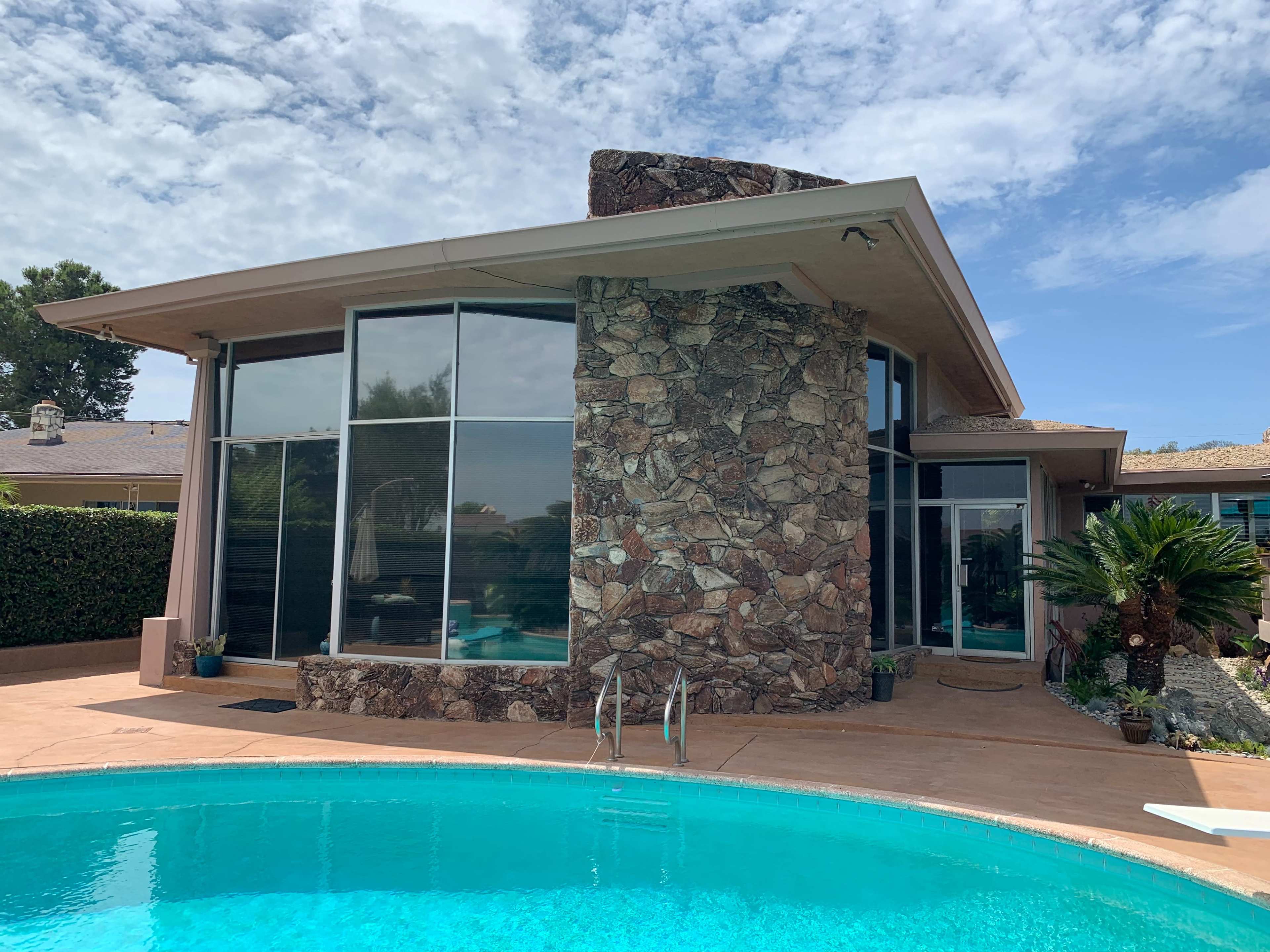 A modern house with a stone feature wall is situated next to a turquoise swimming pool under a partly cloudy sky.