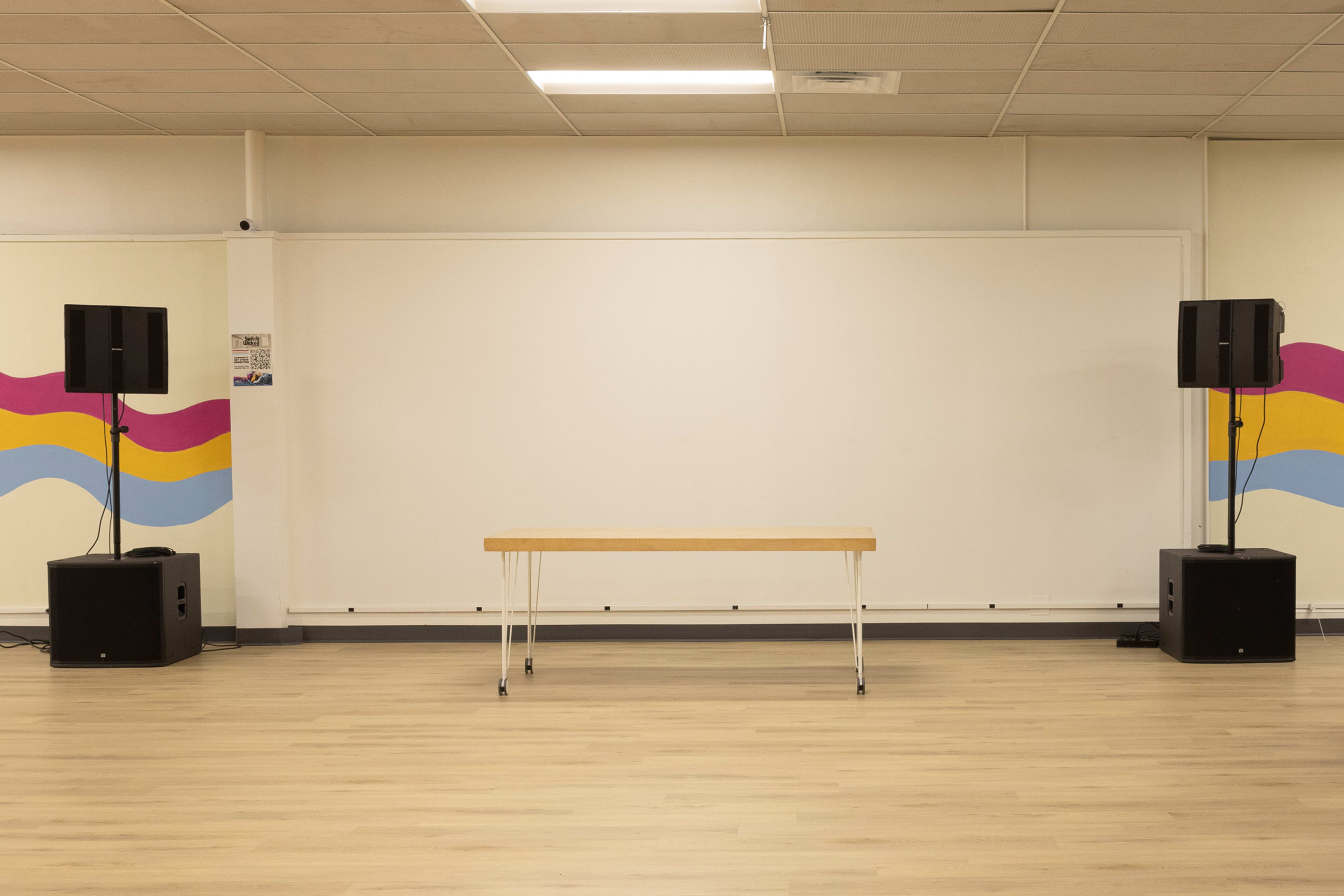 The image shows a spacious room with a wooden table in the center, flanked by two large speakers, and a white wall featuring colorful wave artwork.