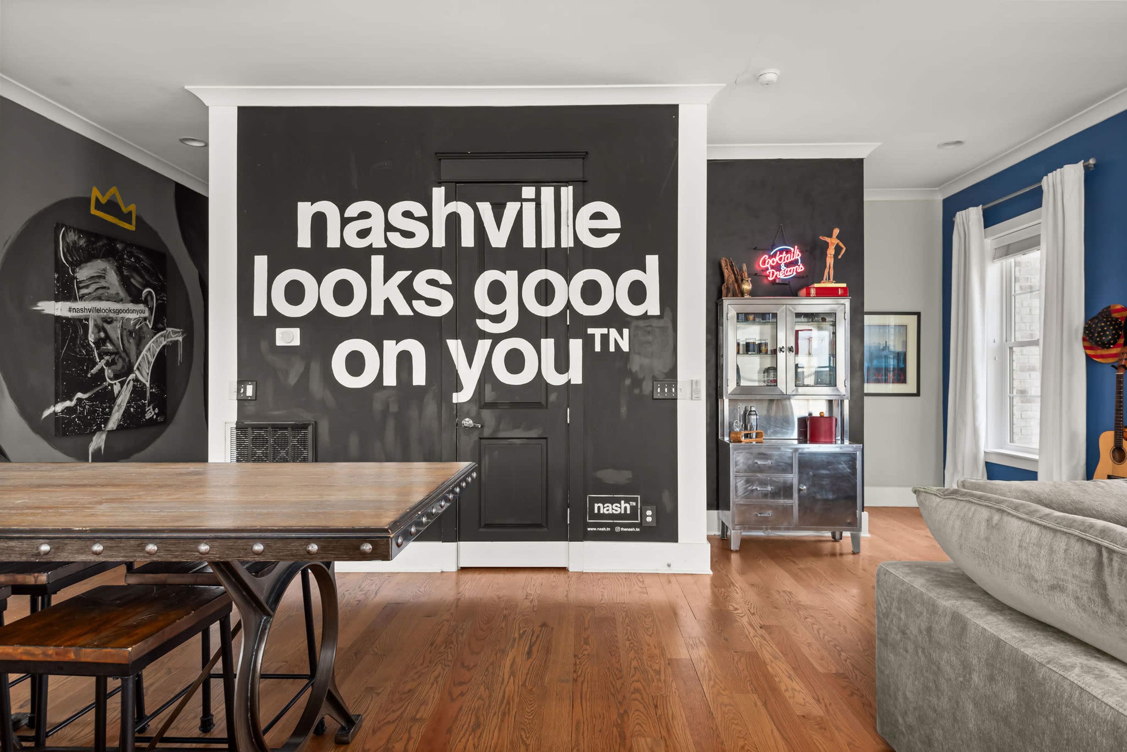 A large black wall featuring the phrase "nashville looks good on you" is positioned behind a dining table in a well-lit room with wooden flooring.