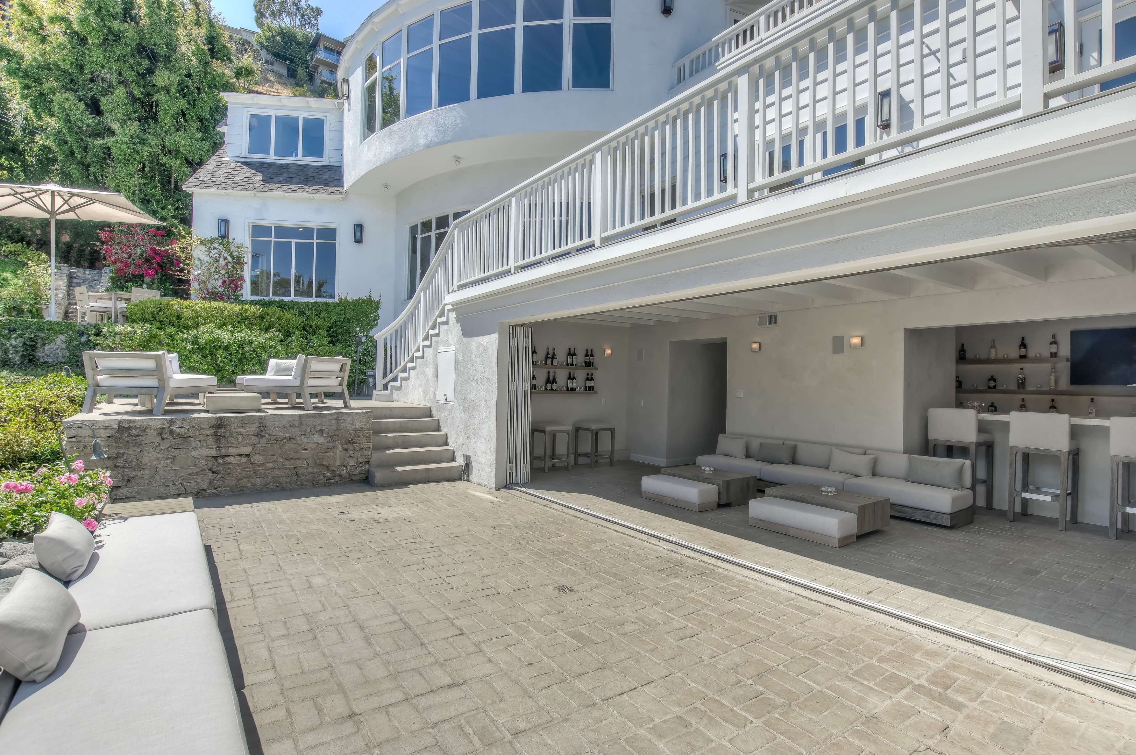 The image shows a modern outdoor patio area of a house, featuring a seating arrangement and a bar area beneath a large balcony.
