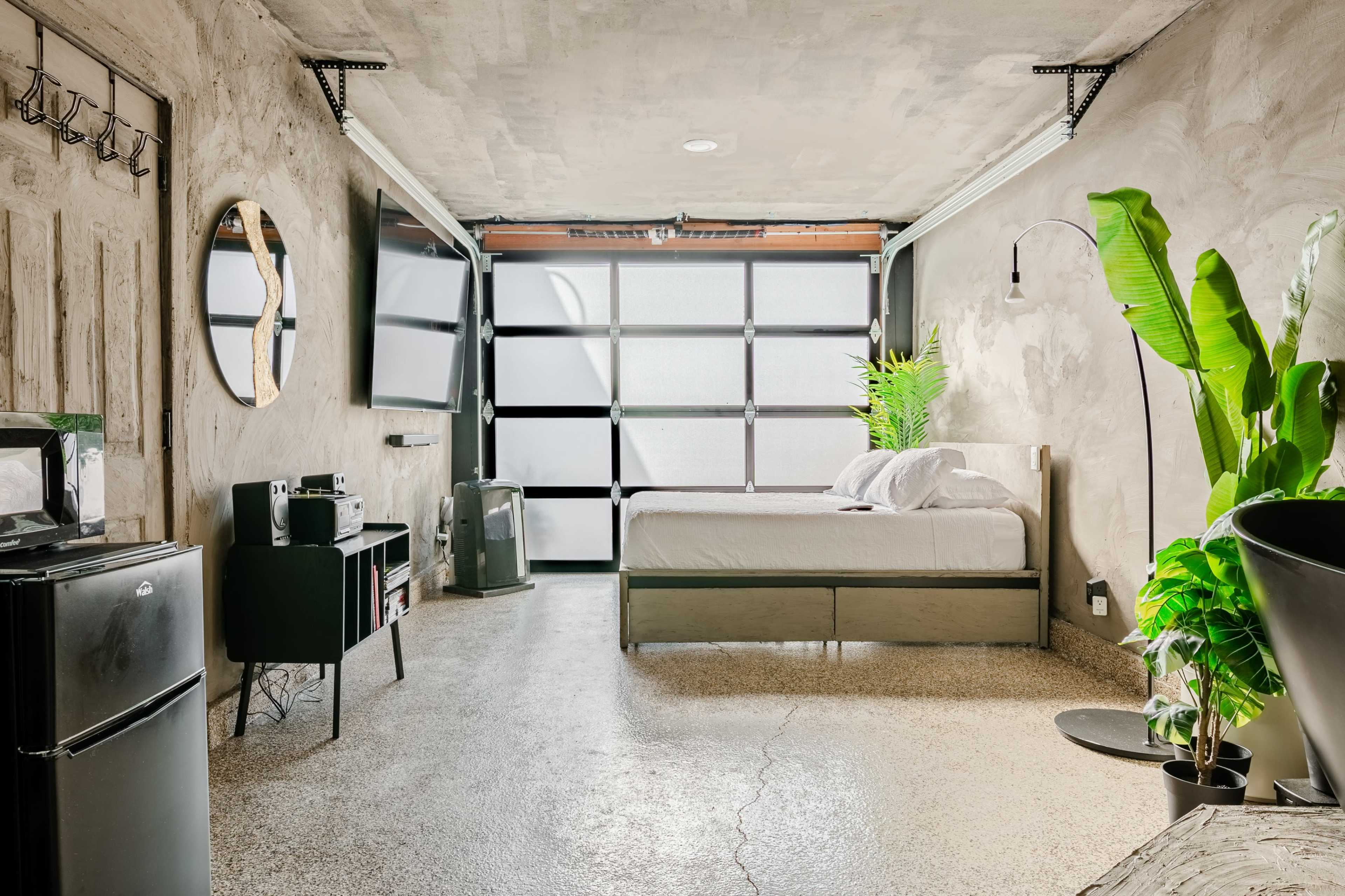 A modern studio apartment features a bed, a television, a small kitchenette, and large windows with concrete walls and polished flooring.