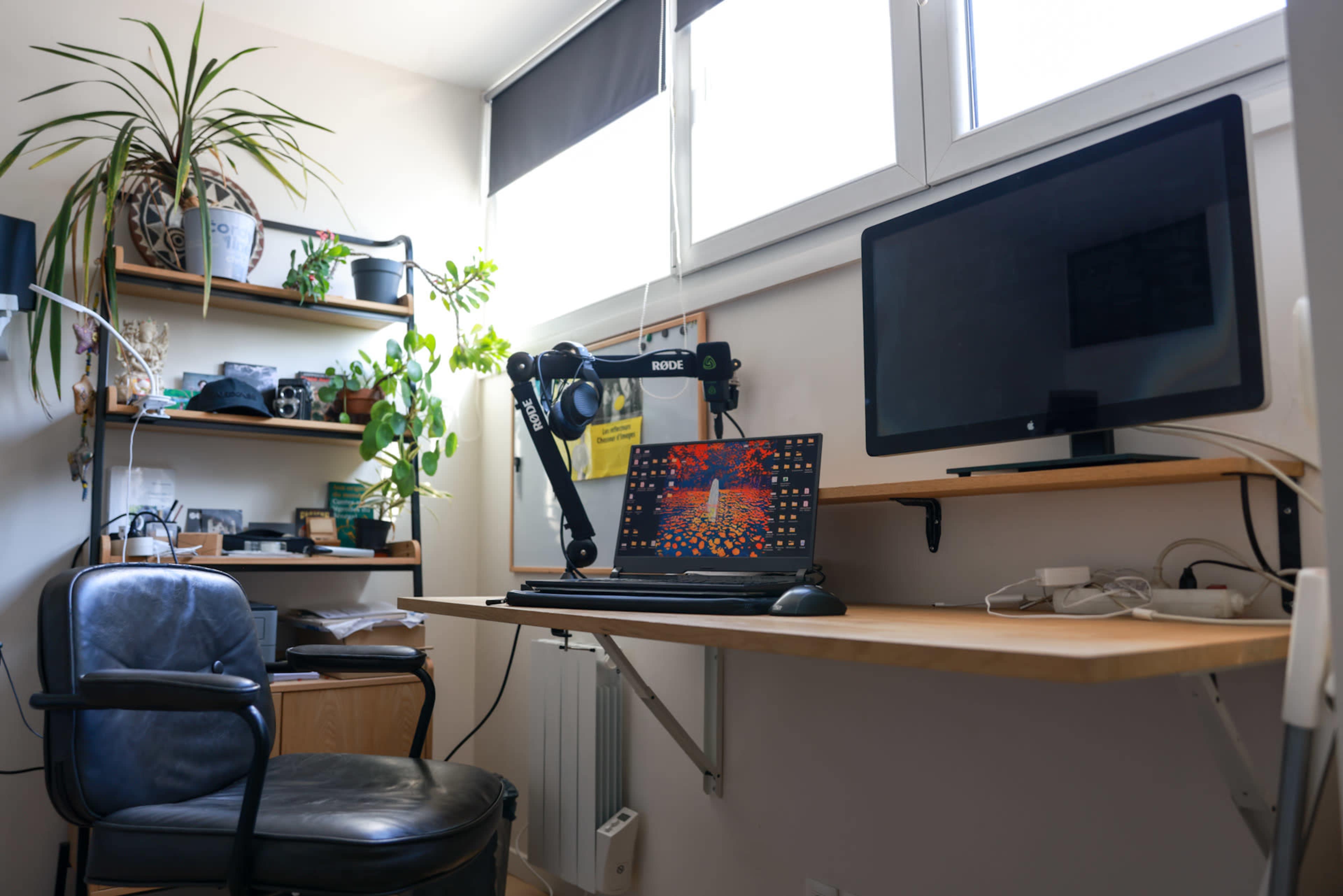 The image shows a workspace with a desk, a laptop, a monitor, a black chair, shelves with various items, and potted plants.