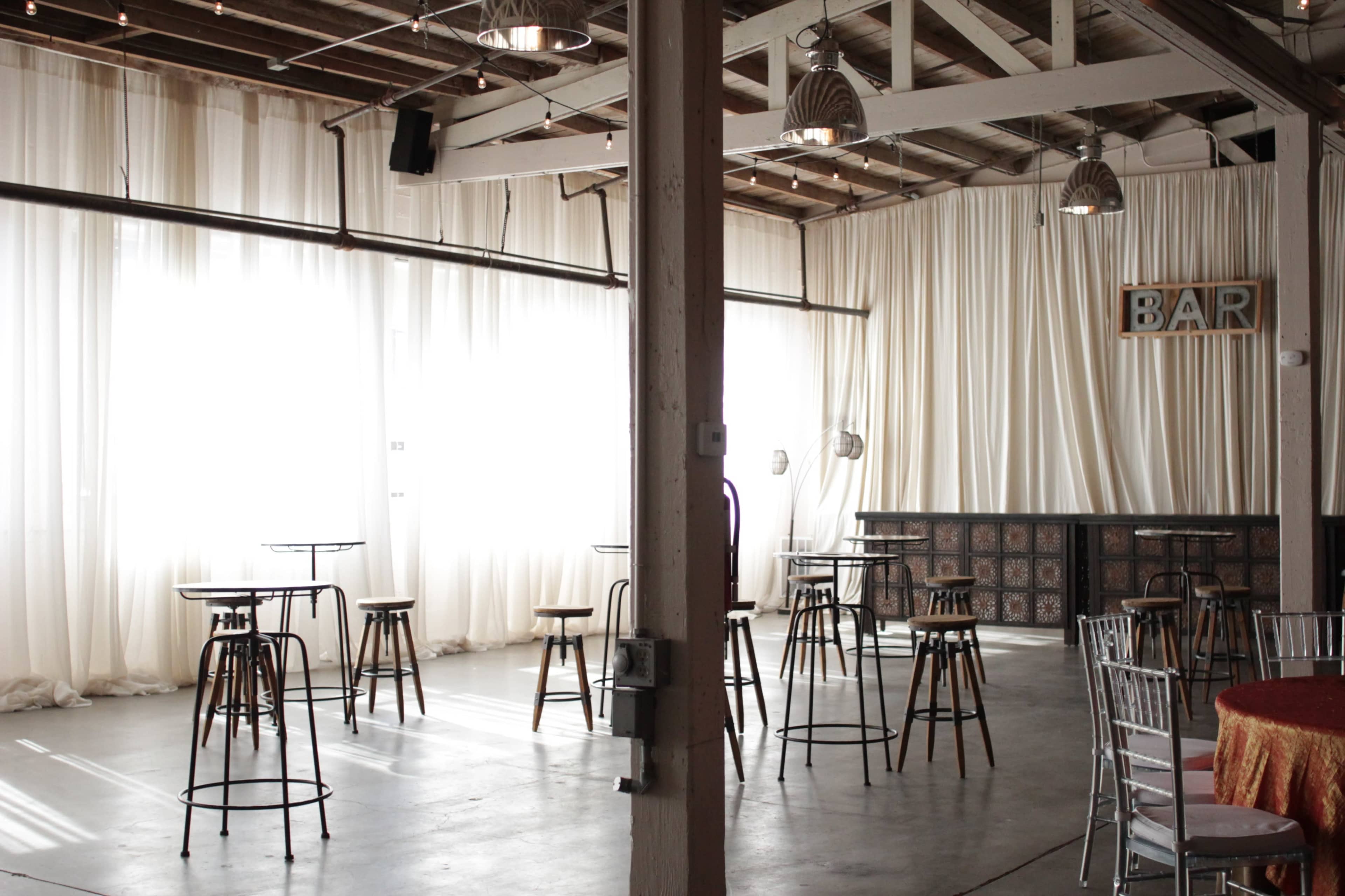 An indoor venue with tall bar tables and stools arranged among large windows draped with sheer curtains, alongside a bar area.
