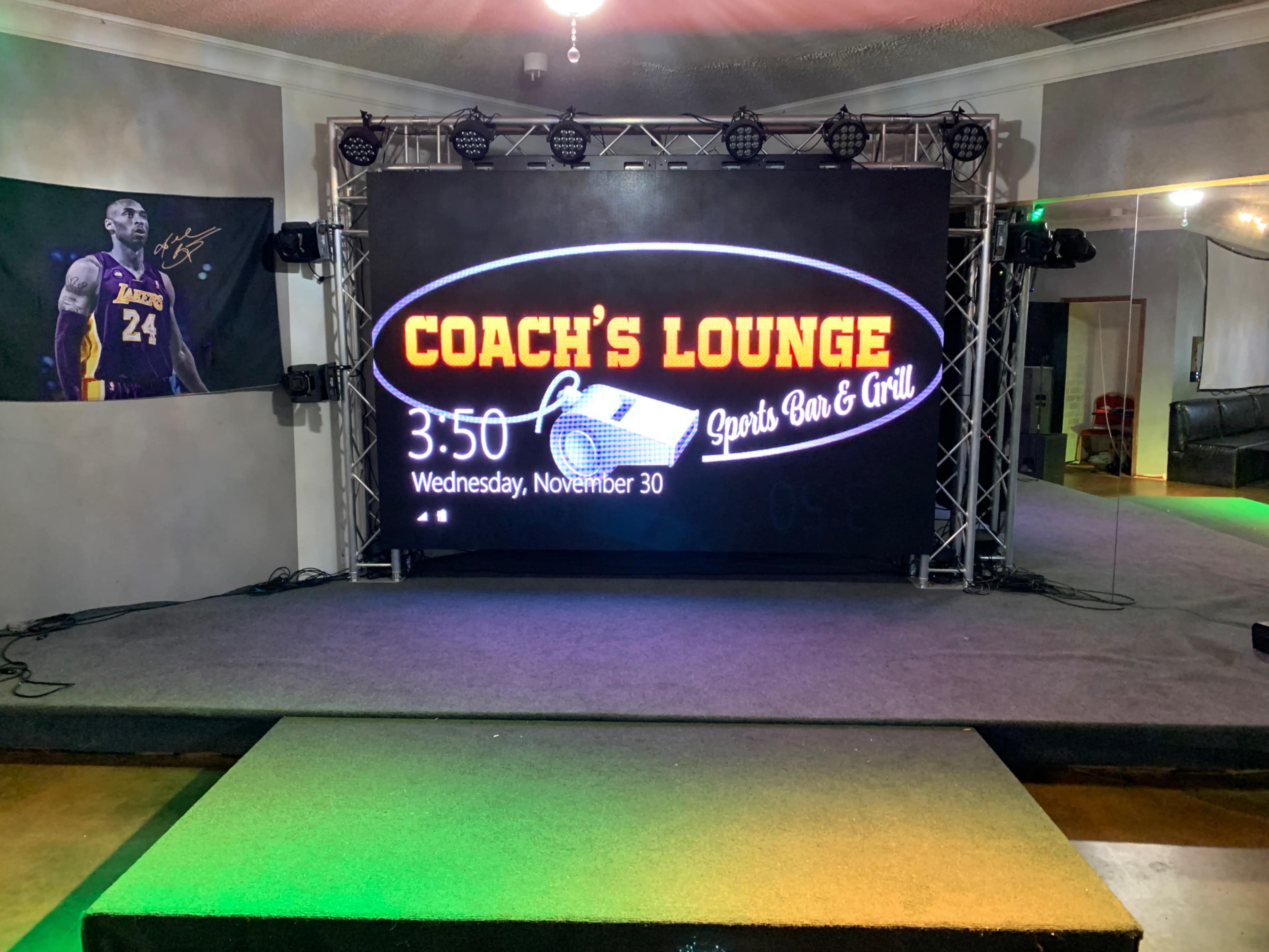 A stage setup at Coach's Lounge, featuring a large digital display with the current time and date, along with a spotlighted area for performances.