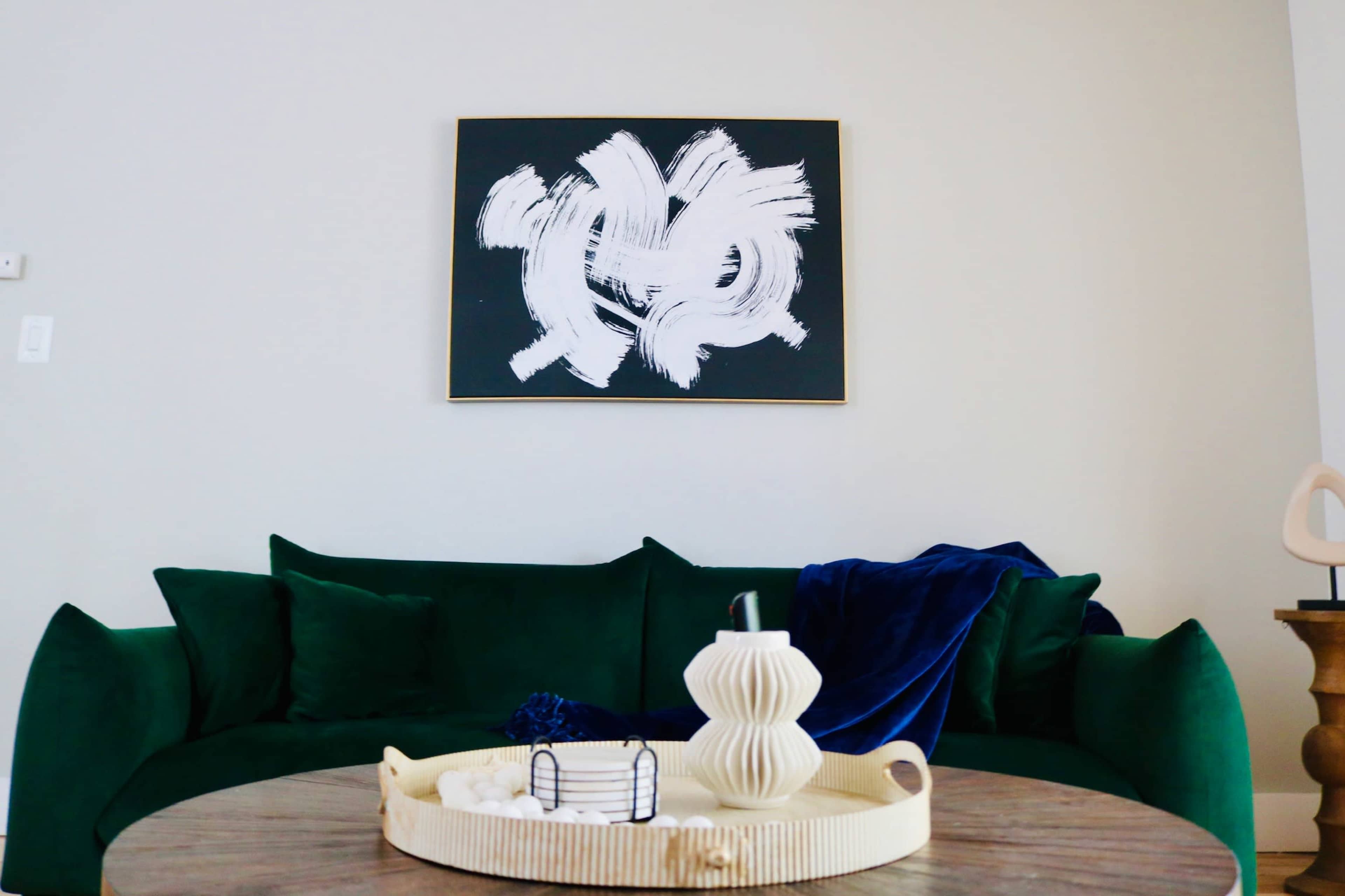 A green sofa is positioned in front of a black and white abstract painting, with a round wooden table topped with decorative items.