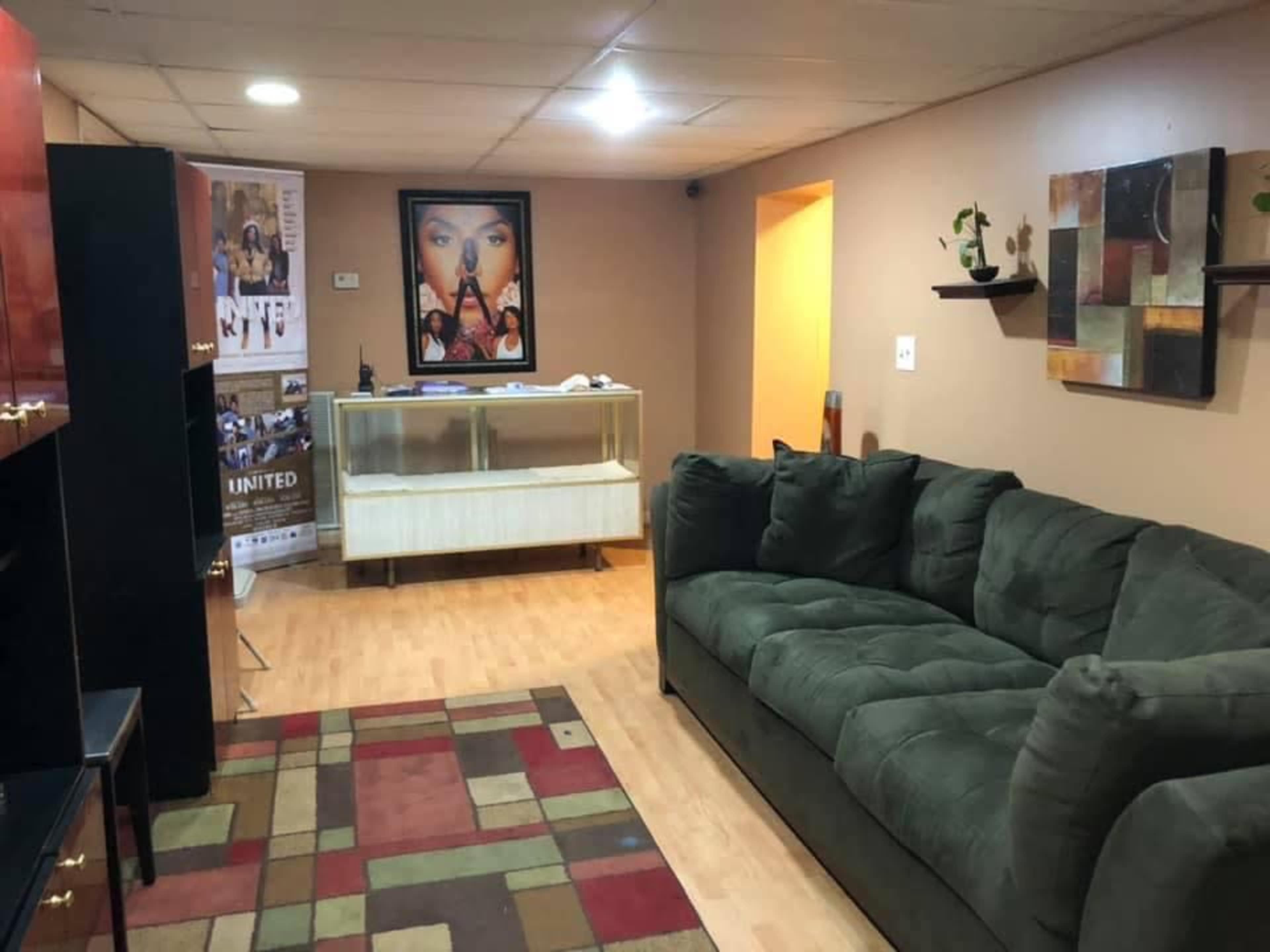 The image shows a cozy room with a green sofa, a geometric rug, a display cabinet, and wall decorations, including posters and plants.