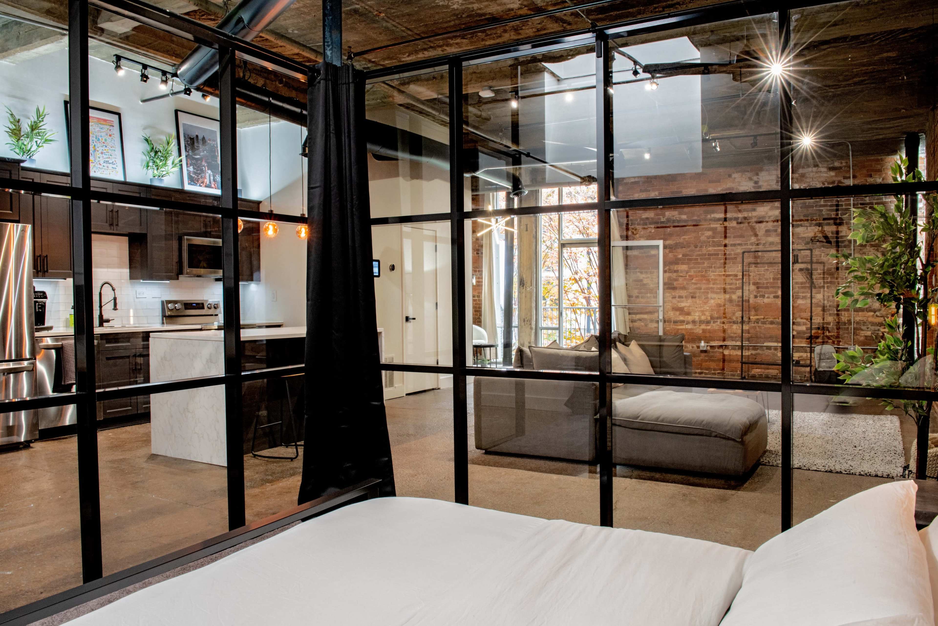 A modern industrial-style room features large glass walls separating a minimalist sleeping area from a living space with exposed brick and natural light.